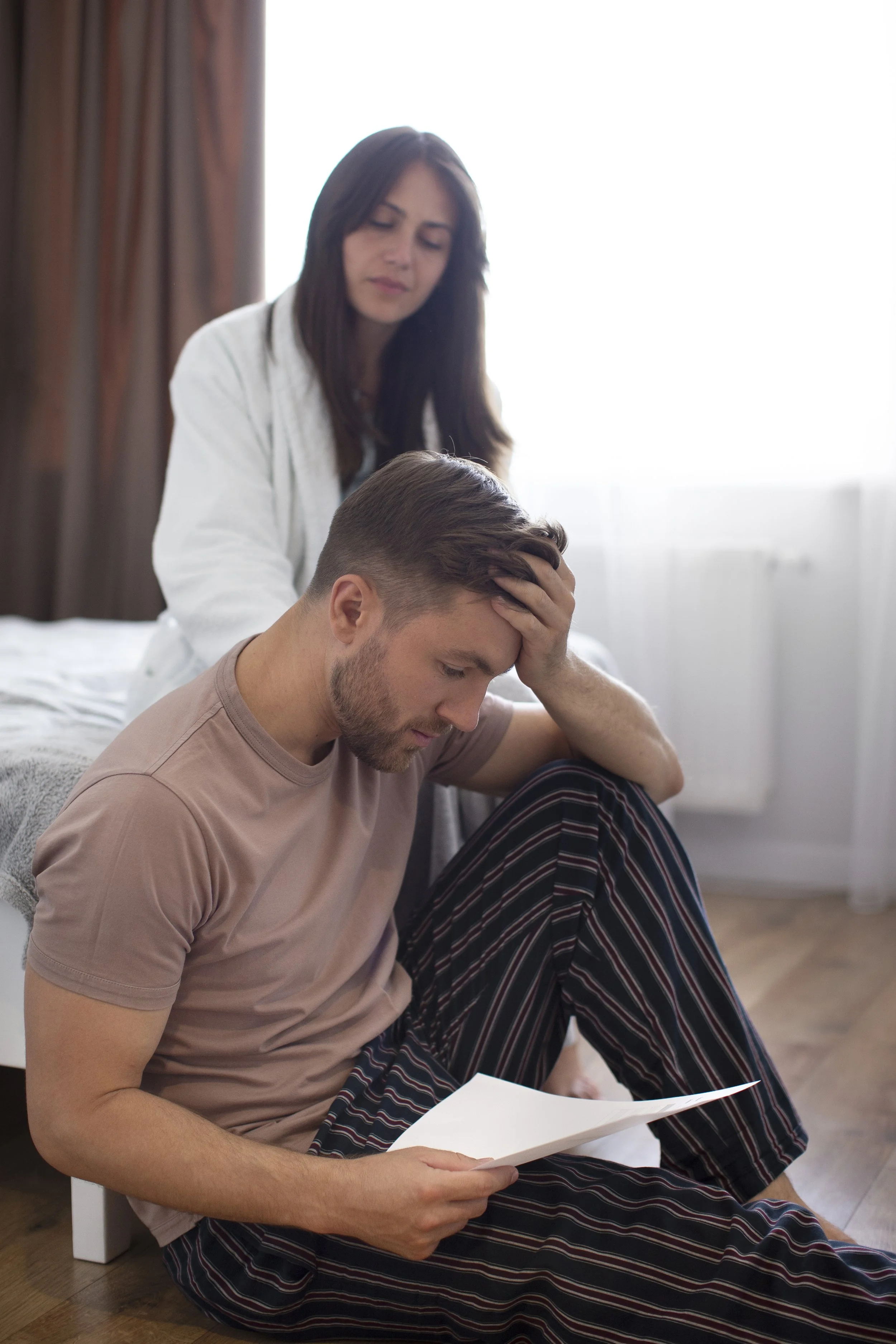 A woman gives a shoulder massage to a man who is sitting on the floor reading a document, both are in a room with natural light.