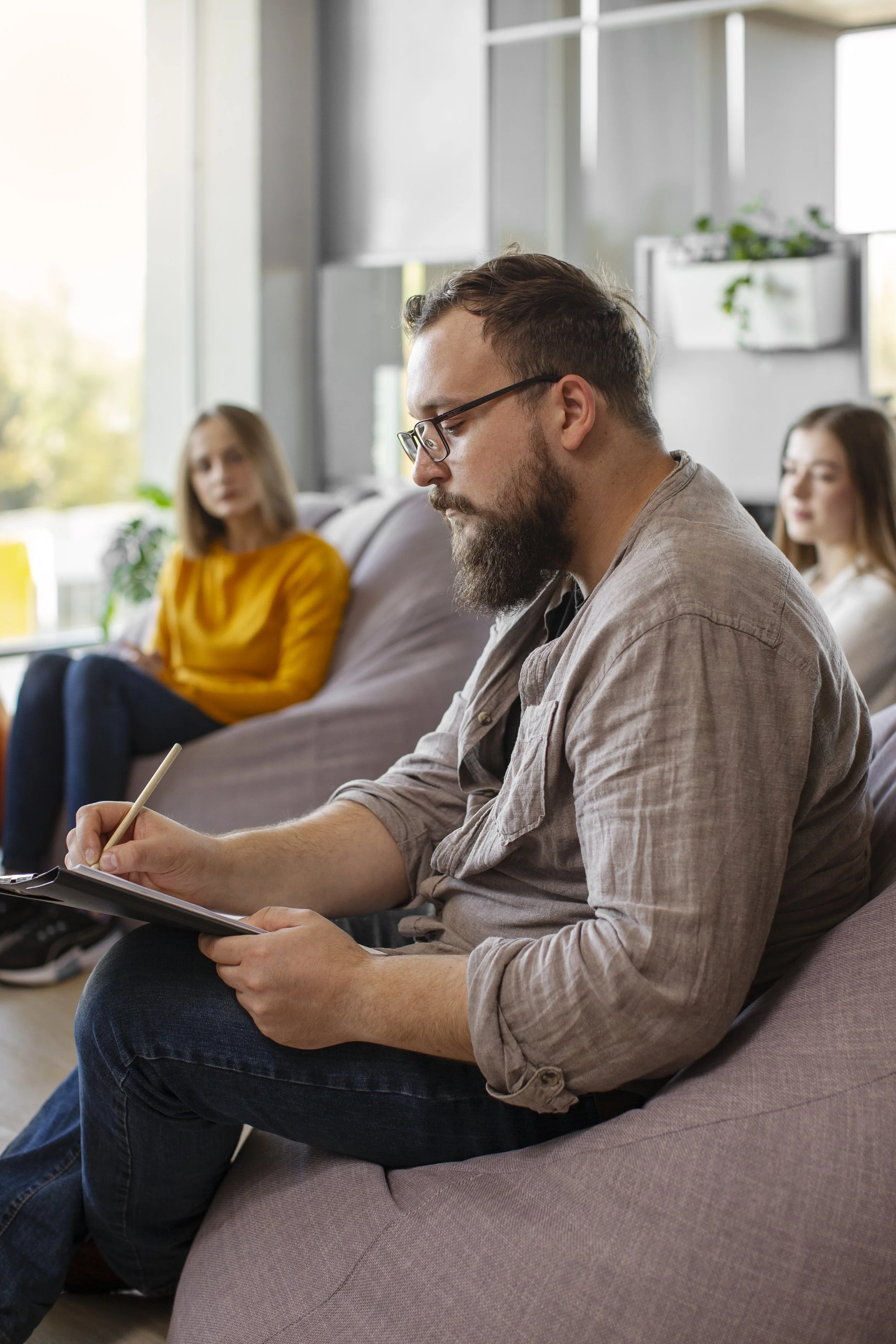 Man with glasses and beard taking notes during a meeting, with two women sitting in the background at a modern office space with large windows and indoor plants.