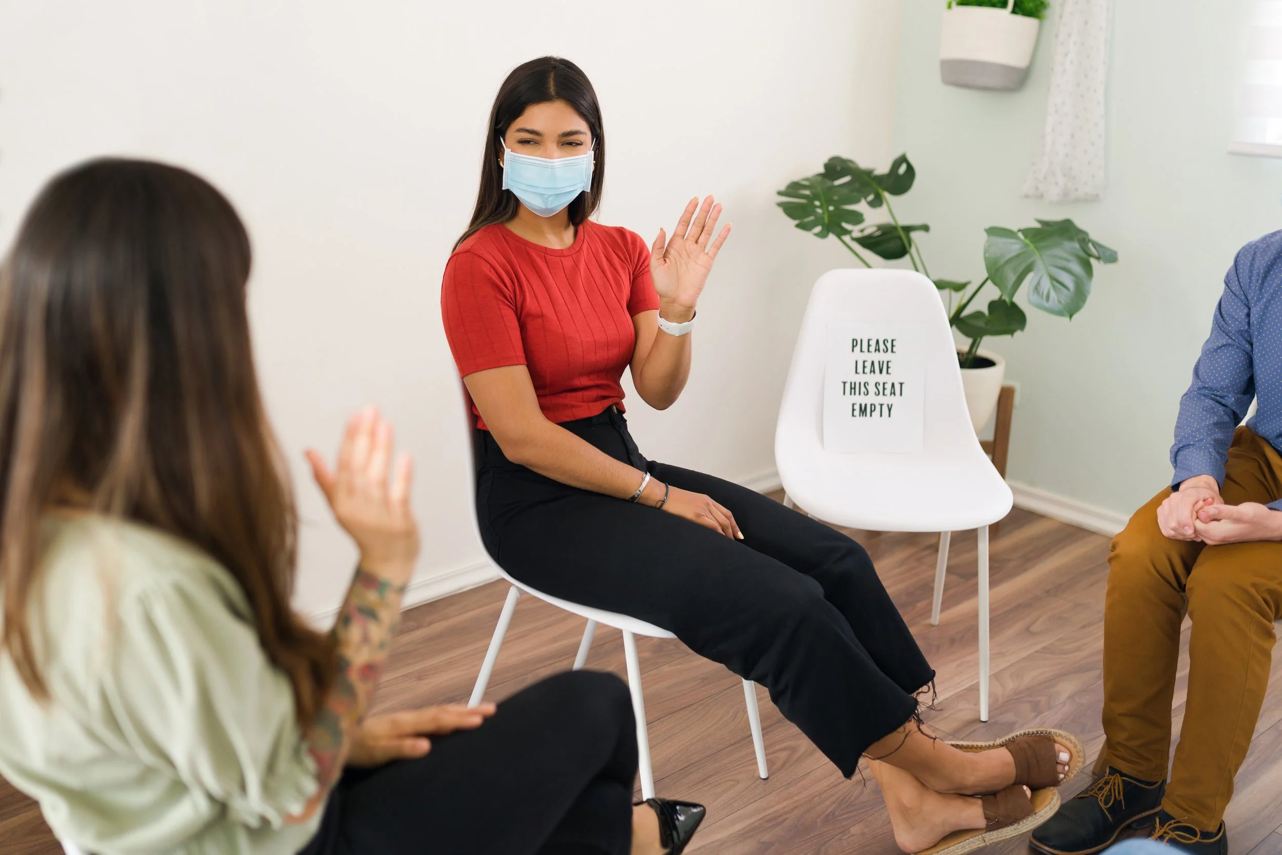 A woman wearing a face mask and red shirt is sitting on a white chair and waving. She is part of a group engaged in conversation in a bright room with a plant and a sign that says 'Please Leave This Seat Empty'.