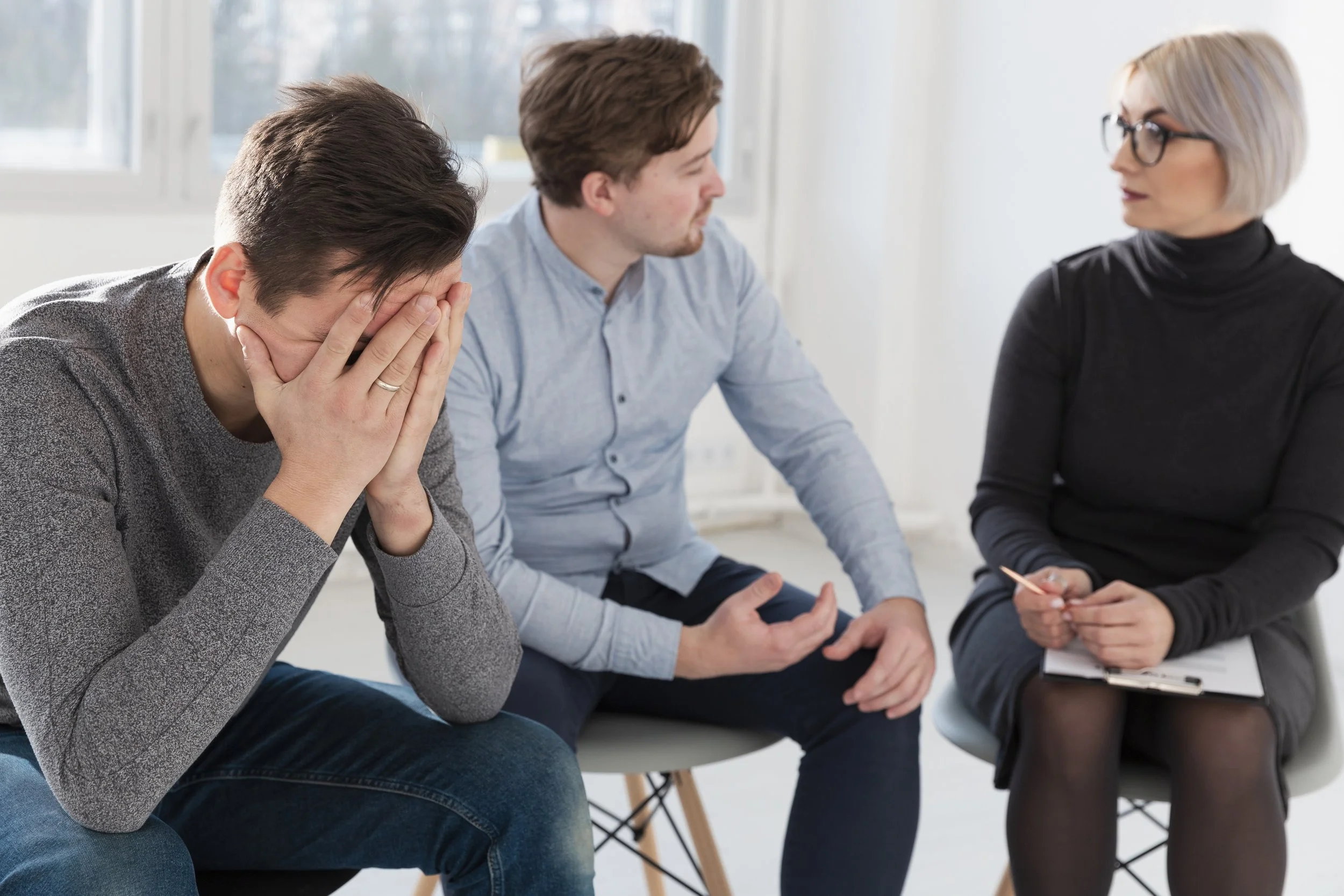 Three people sitting in a room, two men and one woman. One man is covering his face with his hands, looking distressed. The man in the middle is talking to the woman, who is holding a pen and notepad, seemingly listening.
