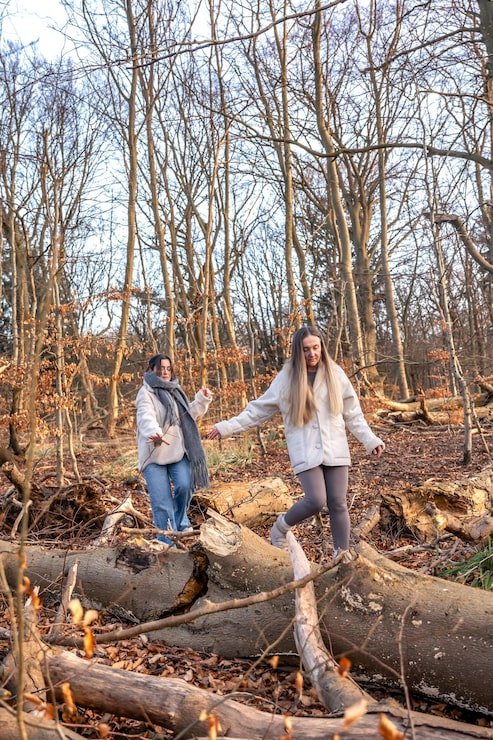 Two women walking on fallen tree logs in a leafless wooded forest during fall or winter.