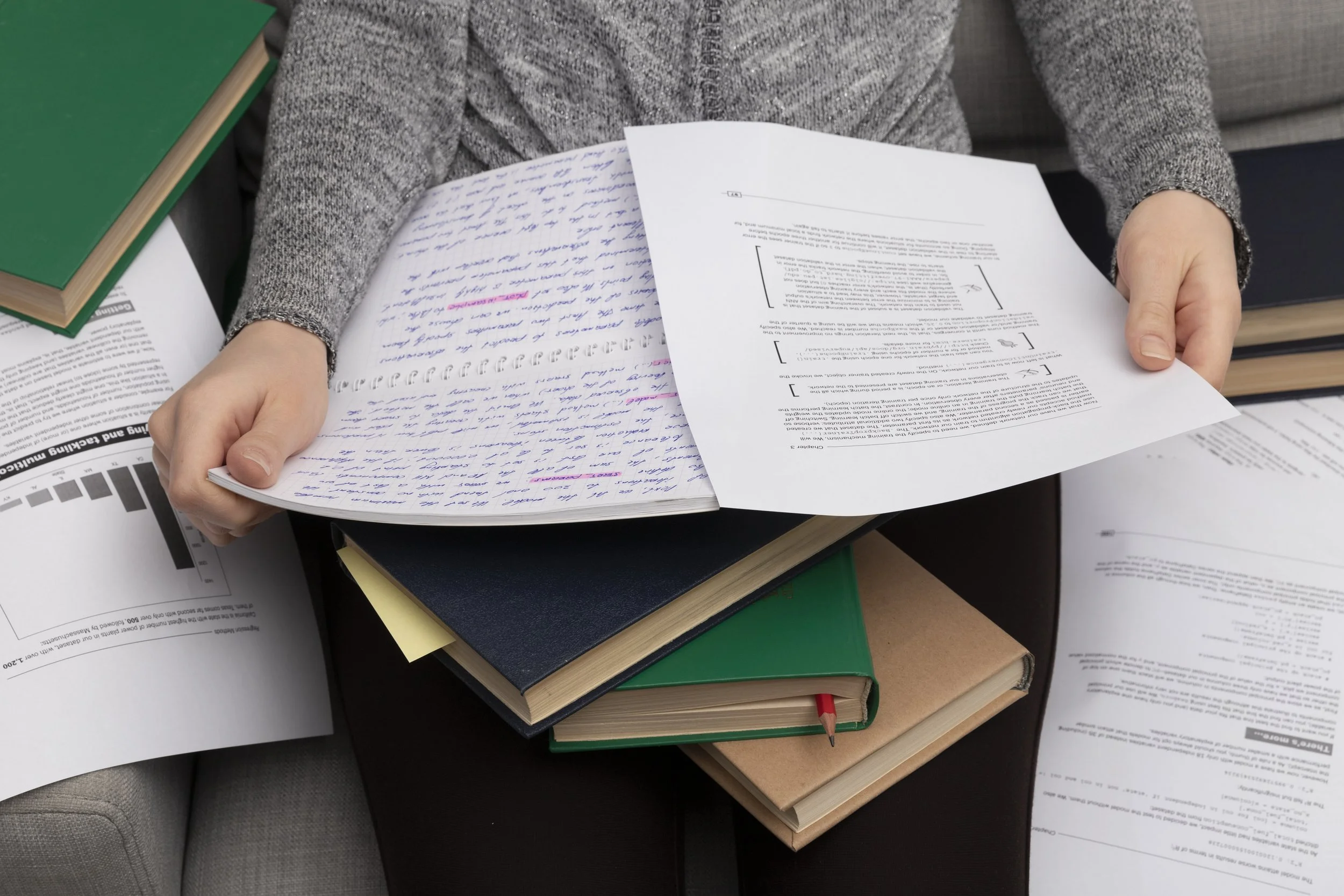 Person holding a printed document with handwritten notes on top of several stacked books on a table.
