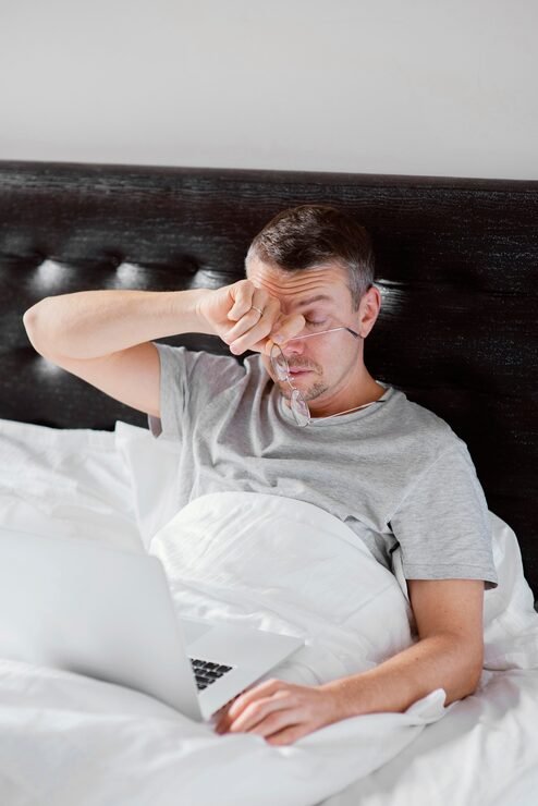 Man in bed with a nasal mask, using a laptop, appears tired or sleepy.