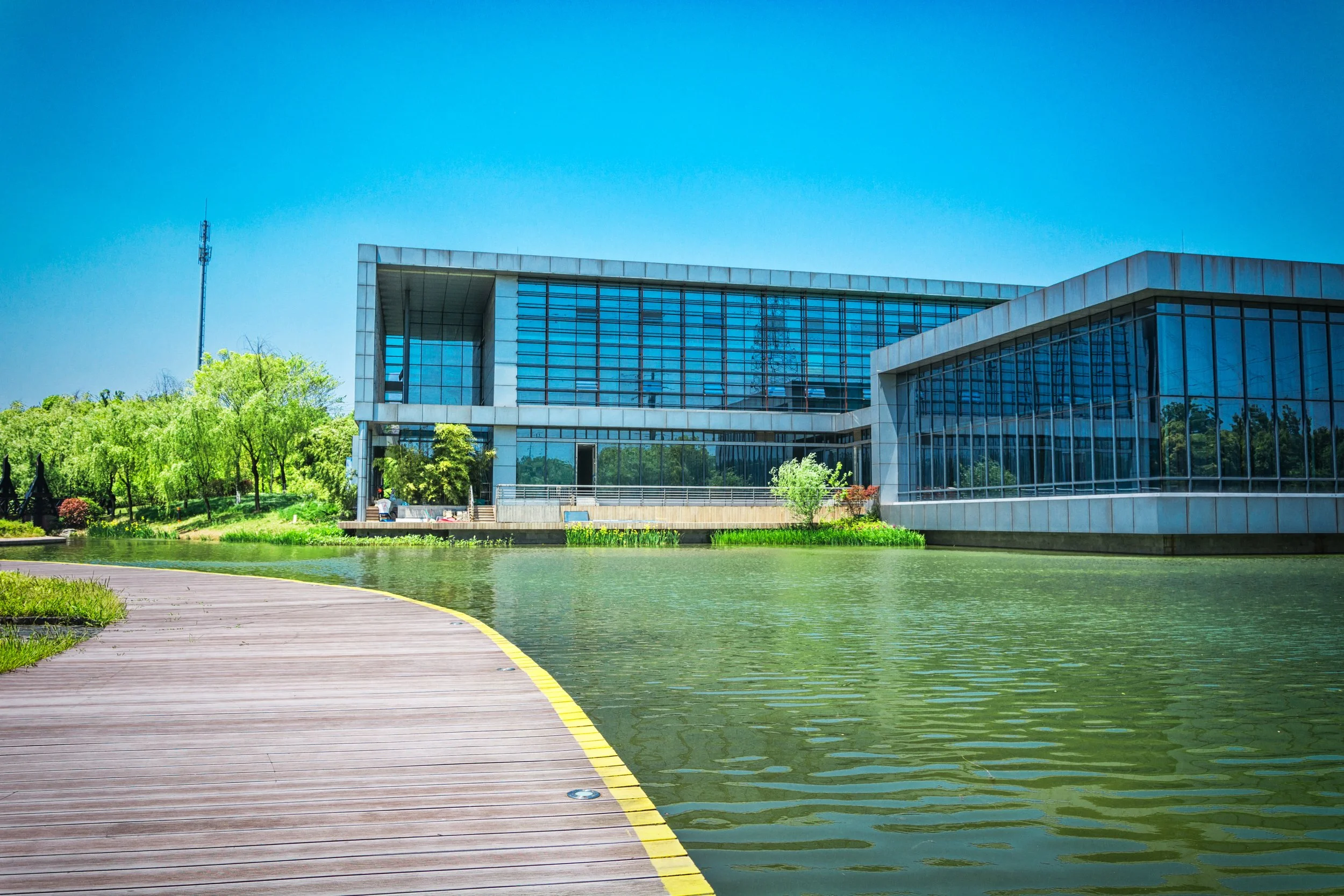 Modern building with glass windows surrounded by trees, a pond, and a wooden walkway under a clear blue sky.