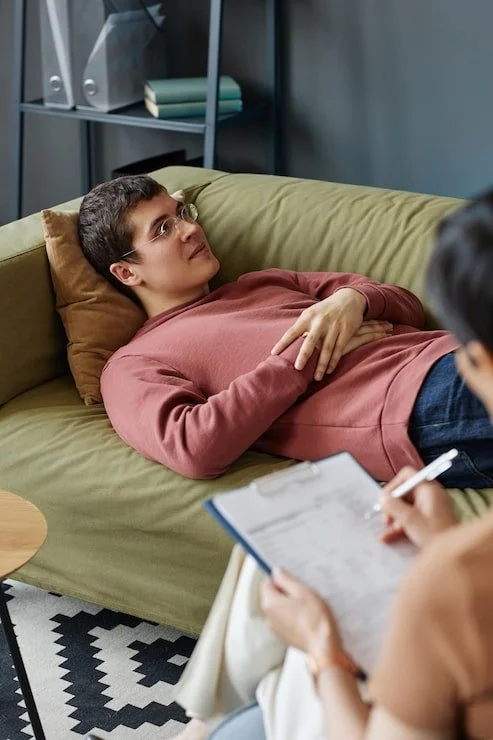 A young man lying on a green sofa in a therapy session, talking to a therapist who is taking notes.