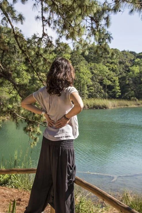 A woman with curly hair, wearing a beige t-shirt and black pants, stands by a lake with her back to the camera, looking out over the water and surrounded by trees.
