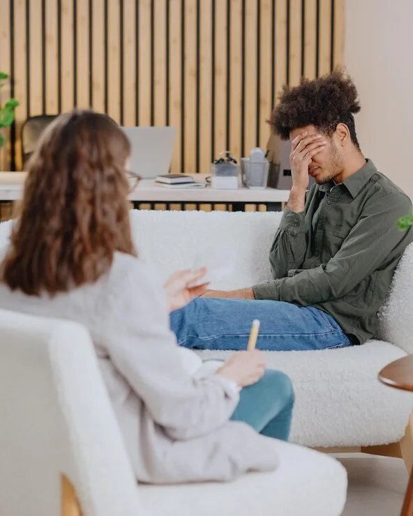 A woman and a man having a therapy session. The woman is taking notes, and the man looks distressed with his hand on his forehead.