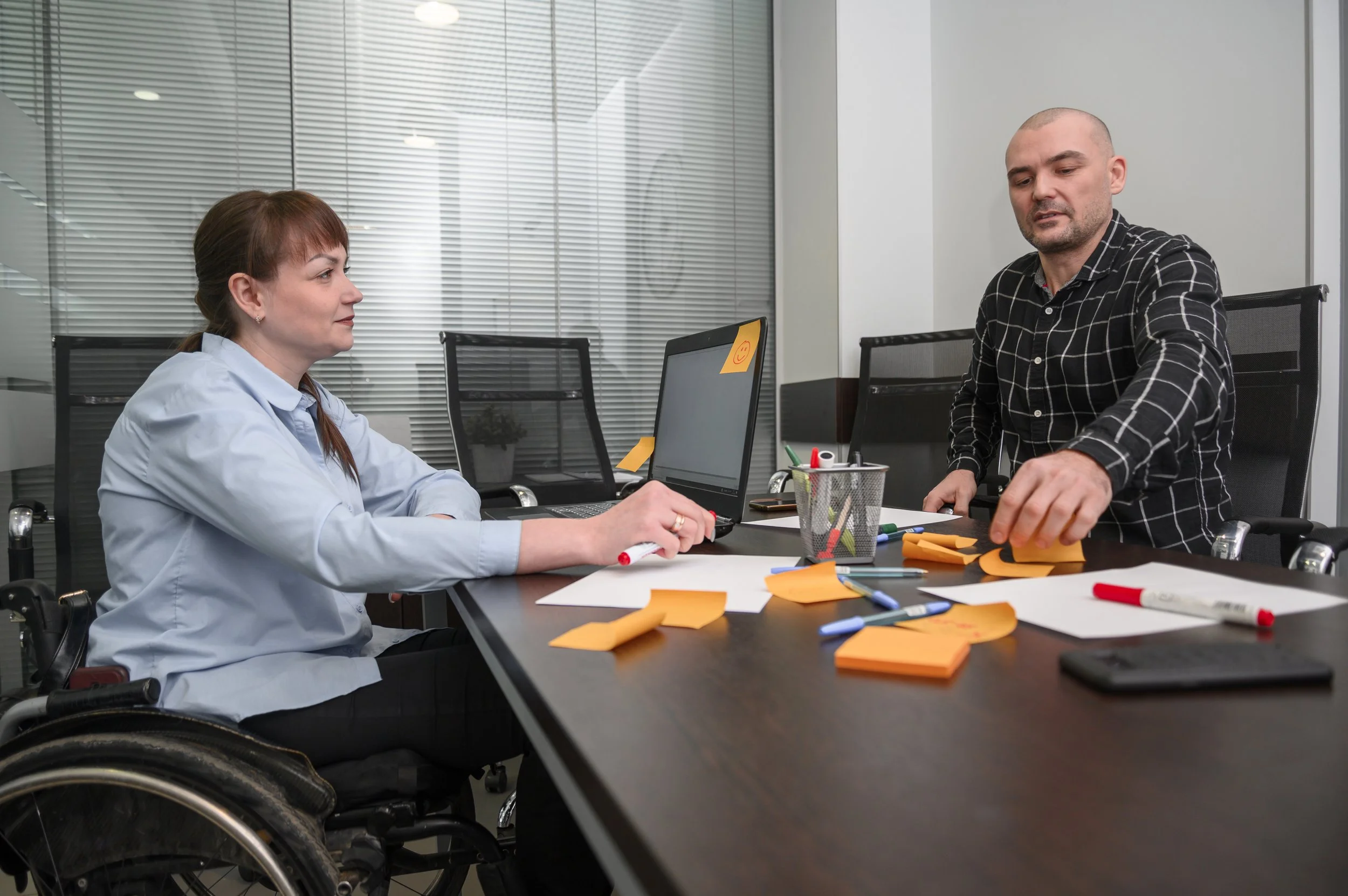 A woman in a wheelchair sitting at a table with scattered orange sticky notes and markers, talking to a man who is standing and reaching for sticky notes in an office setting with glass blinds and desktop items.