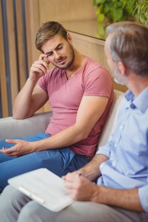 A young man sitting on a couch, holding his wrist with a puzzled or uncomfortable expression, possibly experiencing pain, while talking to an older man who is holding a notepad or clipboard, likely in a medical or counseling setting.
