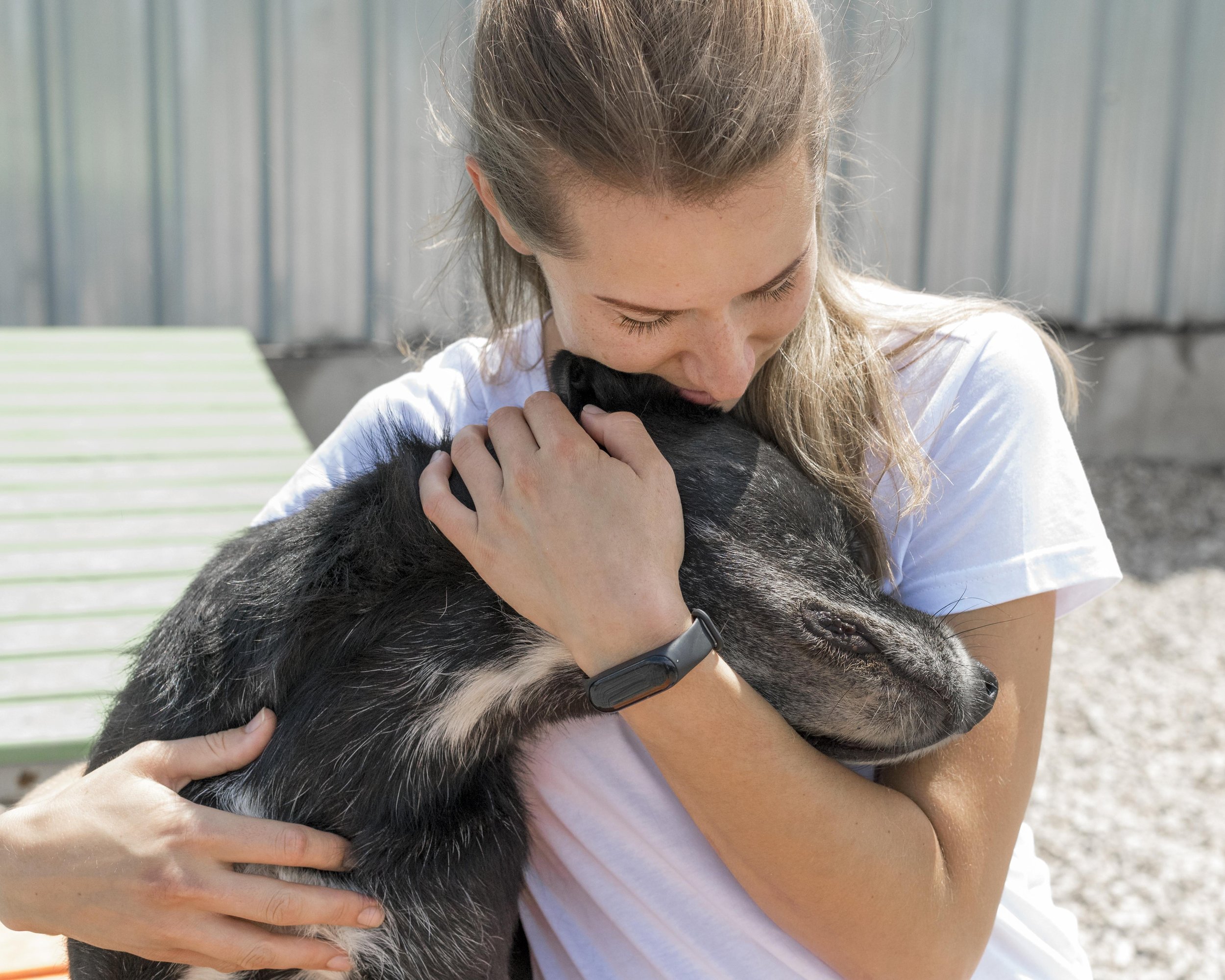 A woman with light brown hair is hugging a black and white puppy, resting her forehead against its head. The woman is outdoors, with a blurred background of metal or wooden panels.