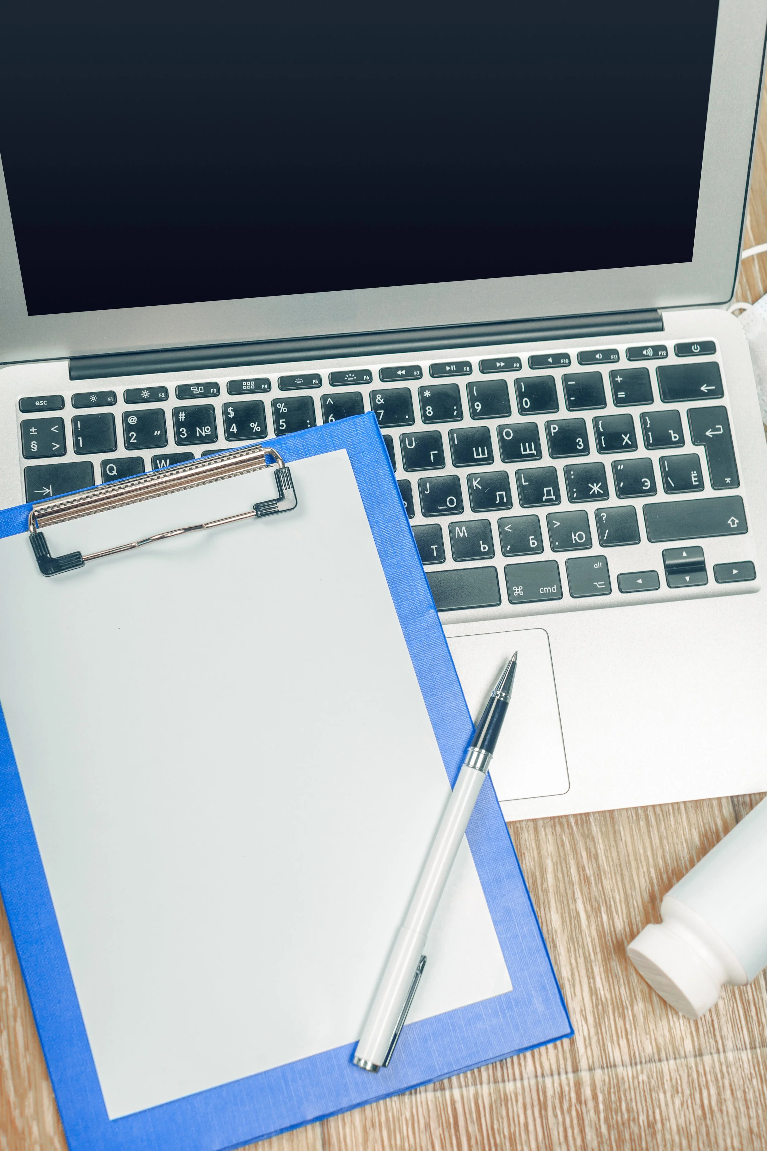 Open laptop computer on a wooden desk, with a blue clipboard, a pen, a white bottle, and a face mask nearby.