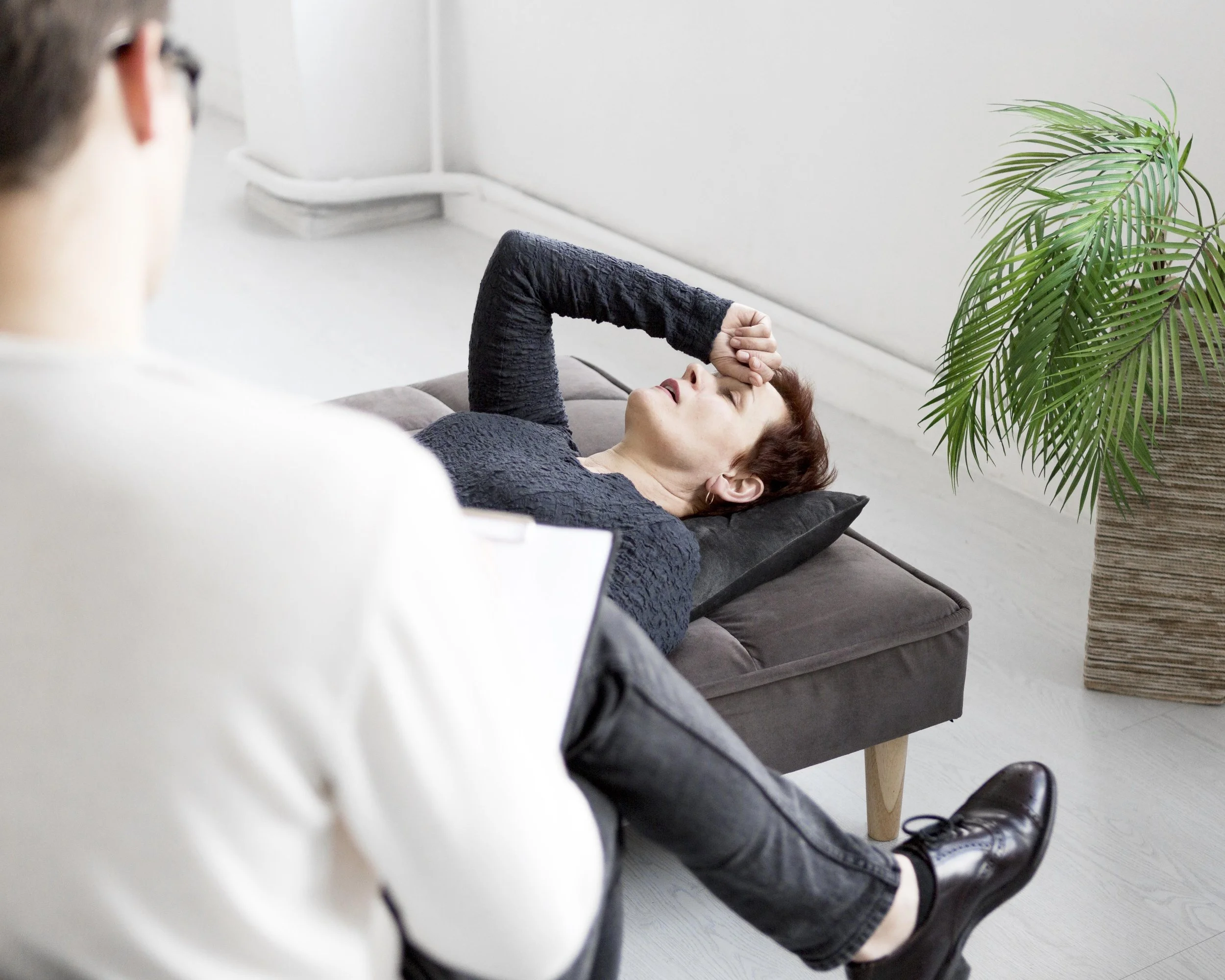 A woman lying on a sofa with her eyes closed and her hand on her forehead, appearing to be resting or possibly tired. There is a person in the foreground, slightly out of focus, and a green plant in a pot beside the sofa.