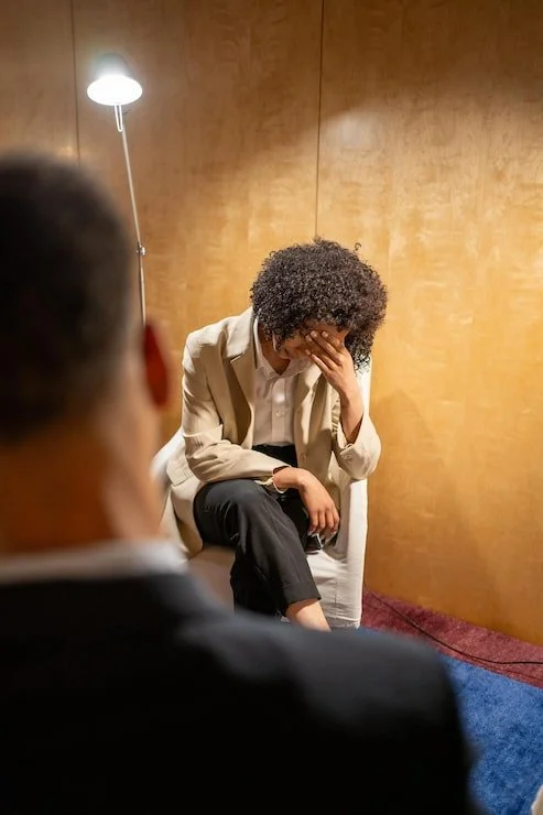 A woman with curly hair sitting on a white chair, covering her face with her hand, appearing upset. She is talking to a person in a suit, seen from behind. The setting is a room with wooden walls and a floor lamp providing light.