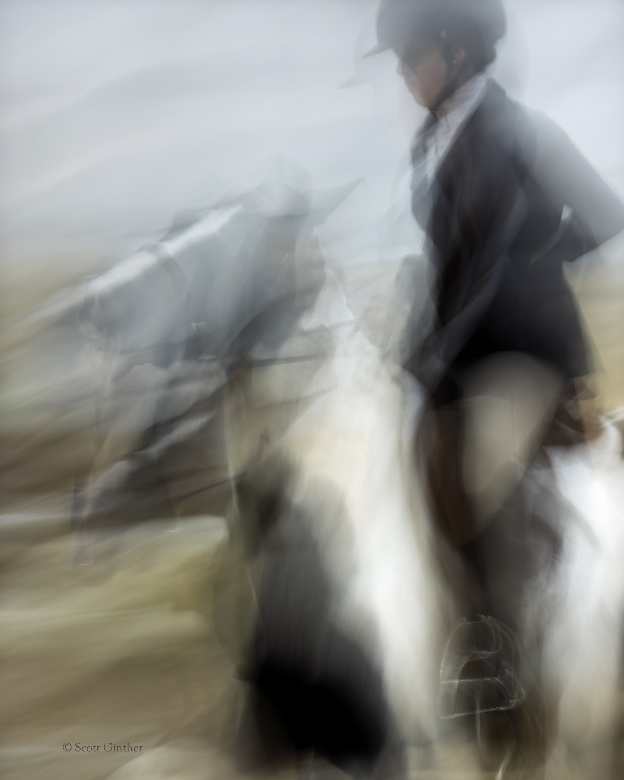 A blurred photograph of a person in an equestrian hat, blazer, pants, and boots is seen from the side. The rider clutches the reins of a black and white horse against a grey sky and brown earth.