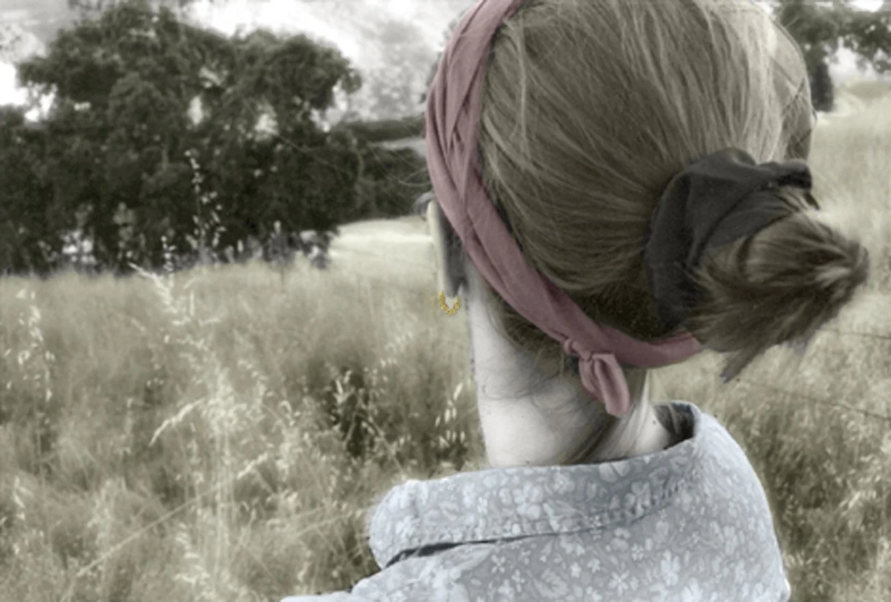 The profile of a woman's head from behind,  her blond hair encircled by a red cloth and secured in a bun. She wears a gold earing and patterned collared shirt.  She stands in a field of tall, yellow grass looking at distant green trees.