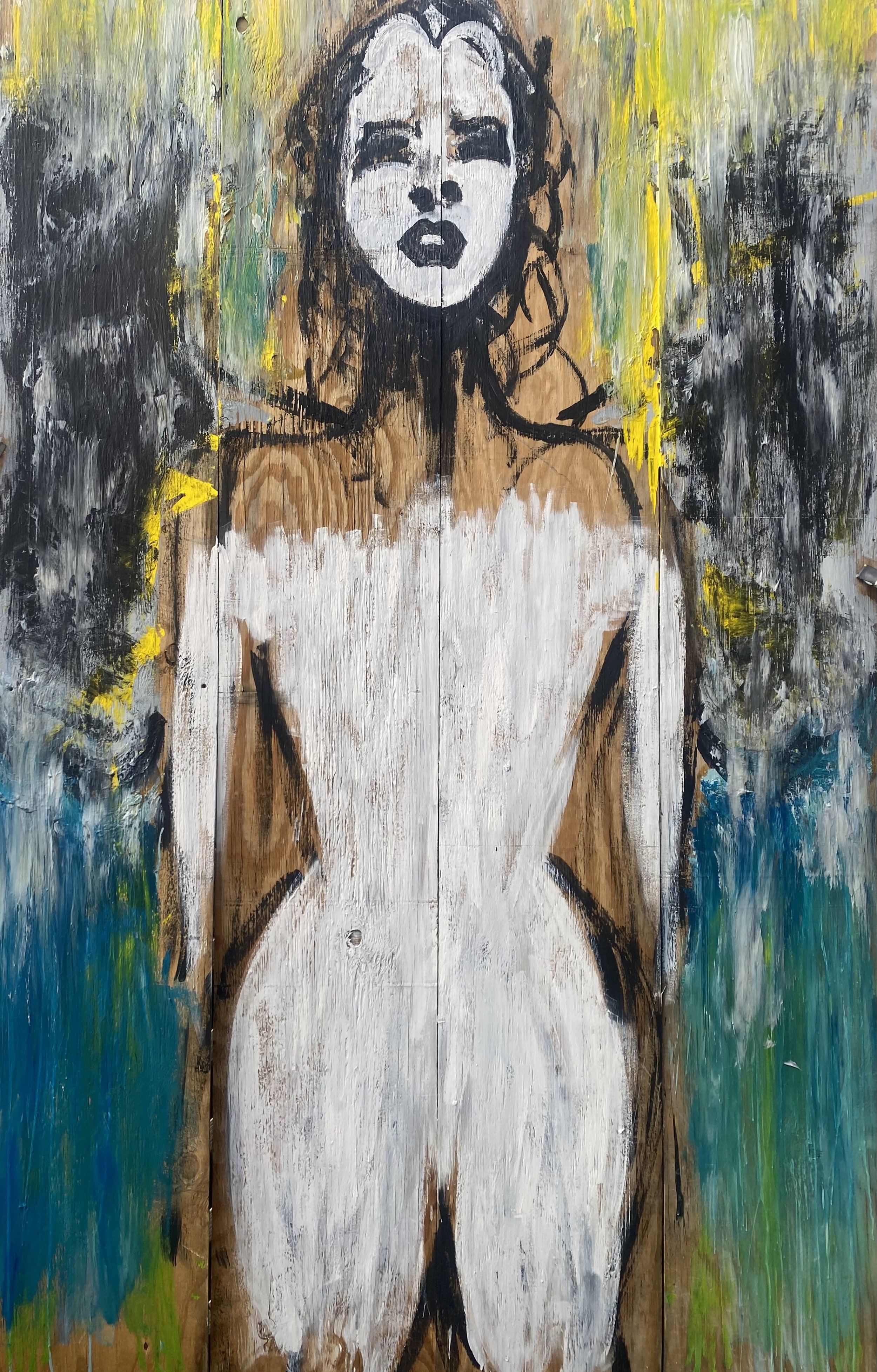 A woman is painted on a wooden board. Her stark white face with black eyes and lips is surrounded by black hair. Her body below the shoulders is the same stark white. The background is streaks of blue, white, green, yellow, black, and white.