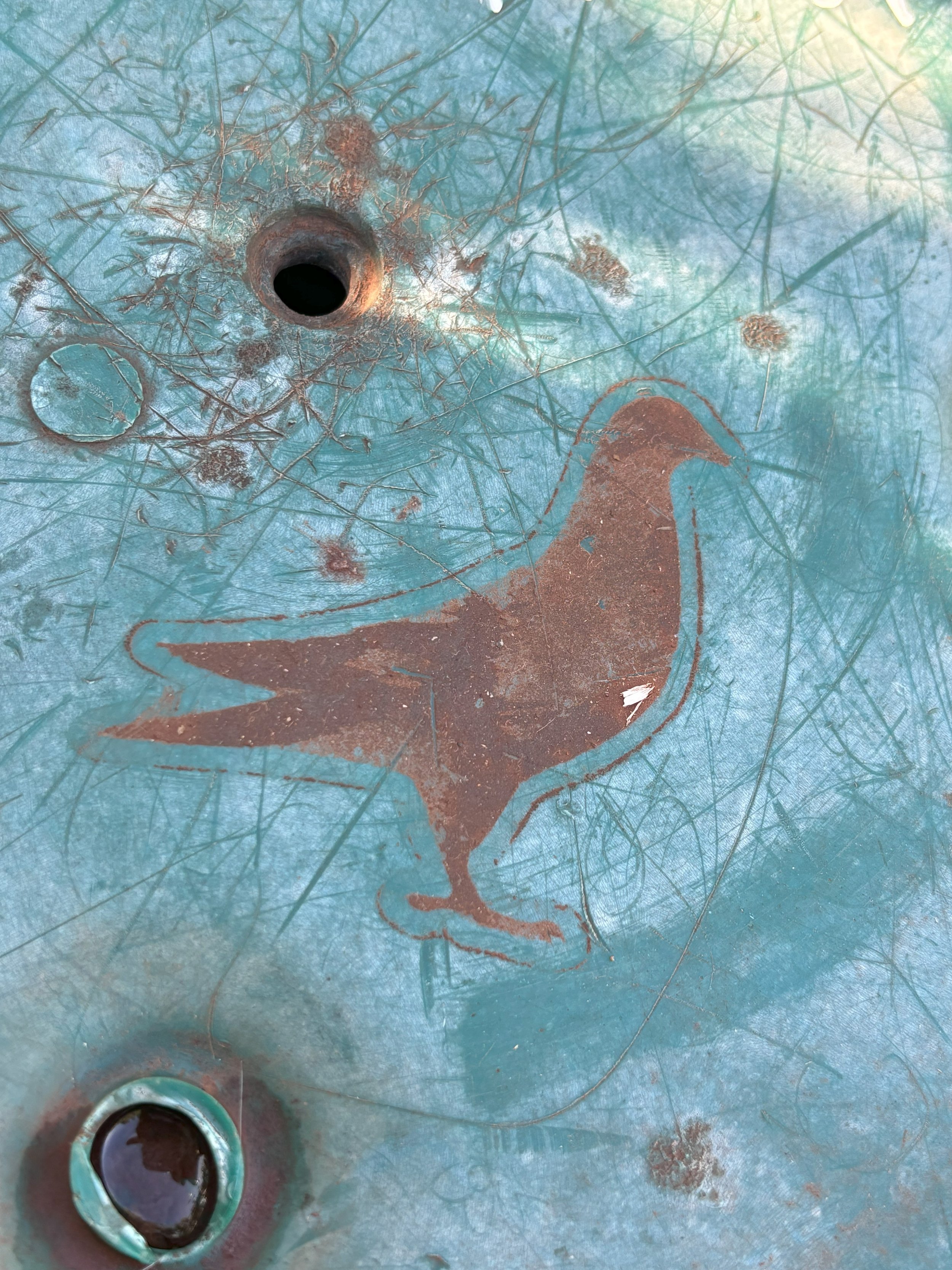 Photo of a blue metal surface with the silhouette of a pigeon and a circular opening above it, both of which are rusted.