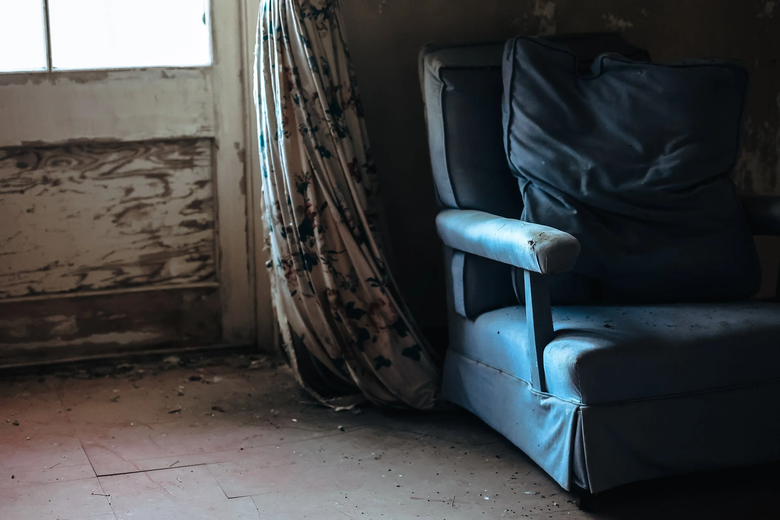 Photo of a dilapidated blue chair partly in the shadows near a window set in a wall with peeling white paint.