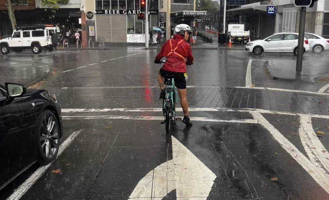 Rider in red clothing and a reflective sash looking over her shoulder while stopped in primary position at Redfern traffic lights in the rain.