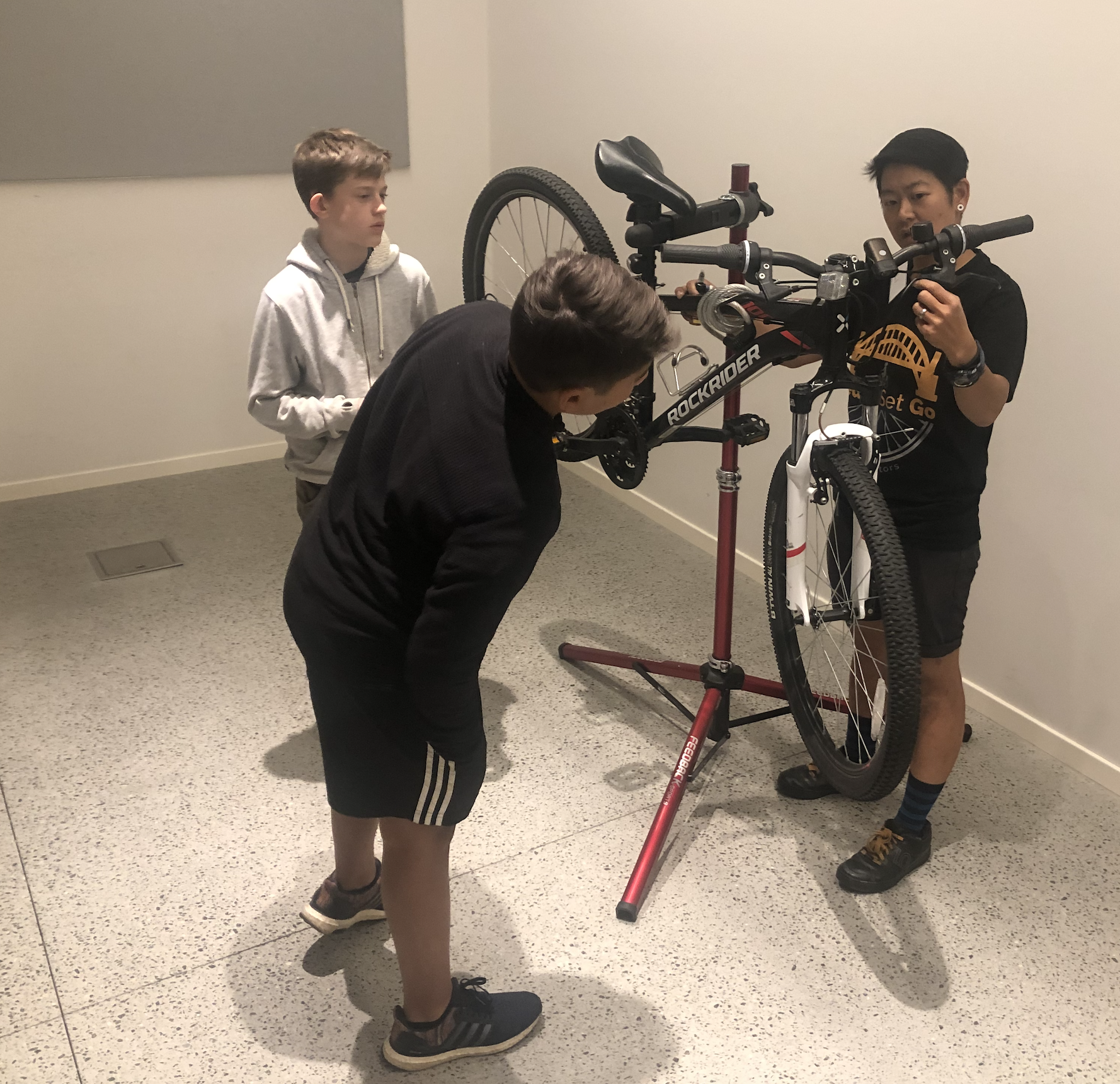 Two teenagers watching a bike maintenance demonstration with an instructor at a repair stand