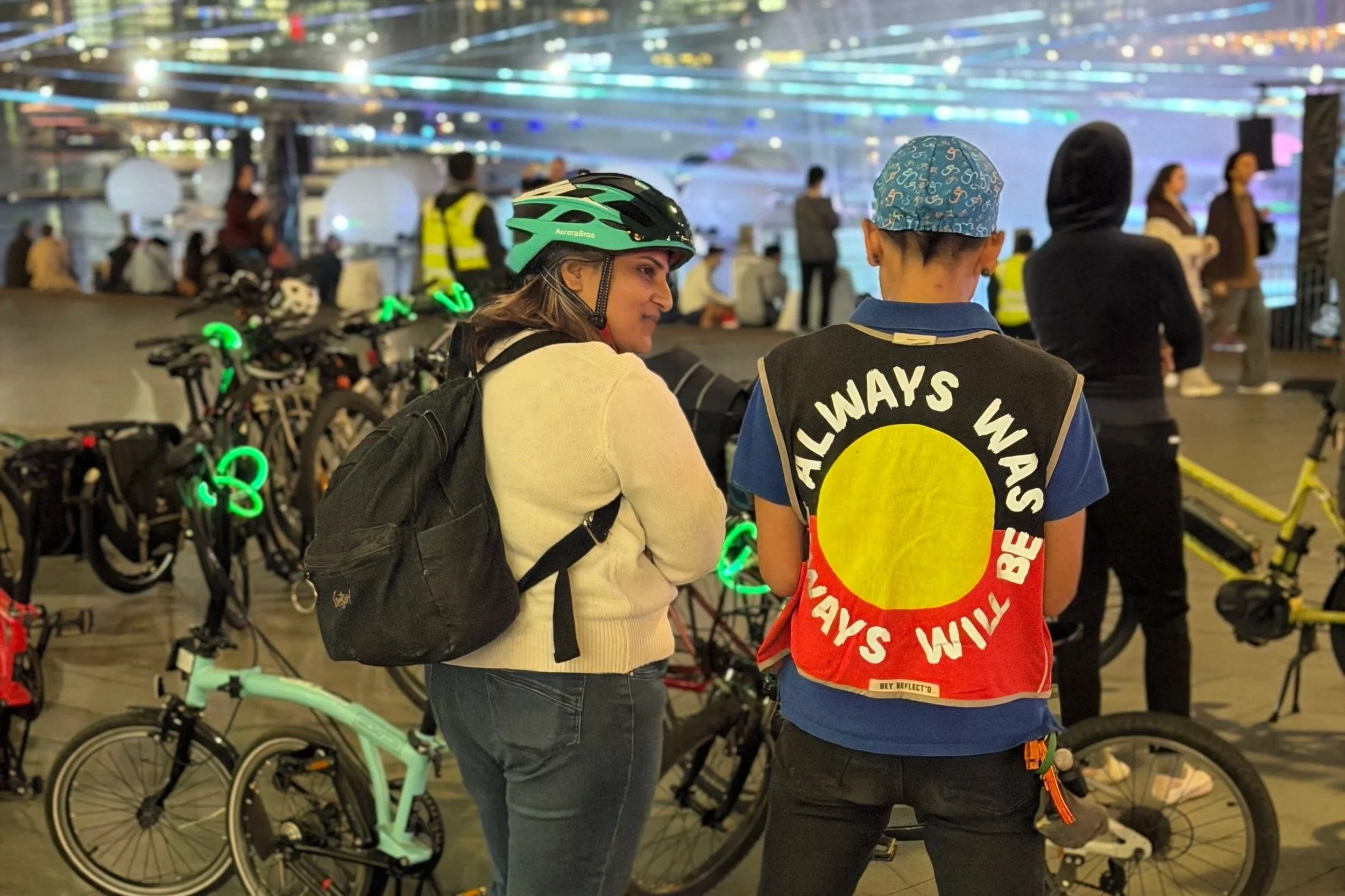 Pedal Set Go guided ride participants with colourful bike lights stopping to enjoy Vivid Sydney at night, with an instructor wearing a reflective Always Was, Always Will Be vest.