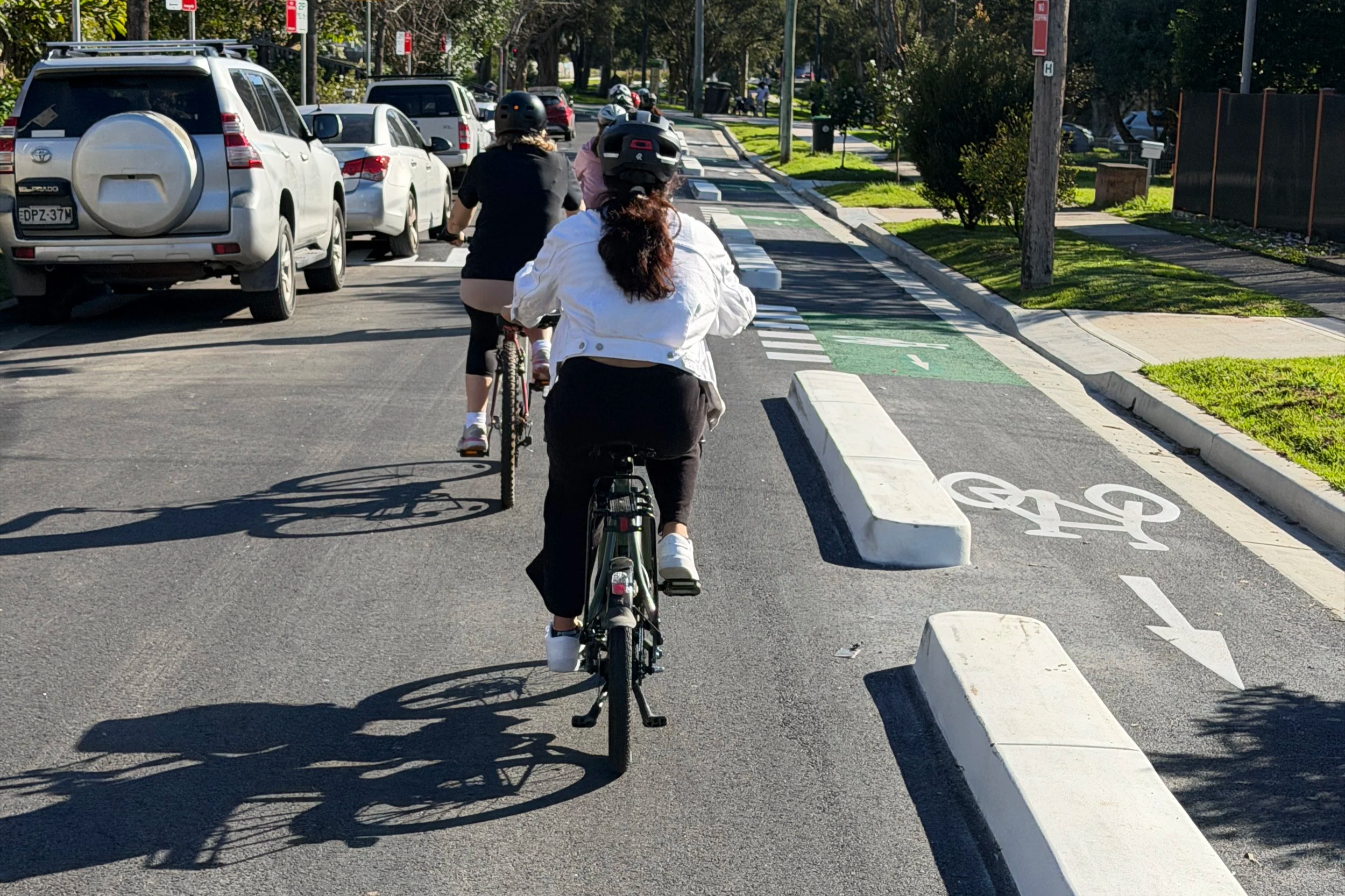 Pedal Set Go group riding single file away from parked cars on a quiet Parramatta street beside a separated cycleway.