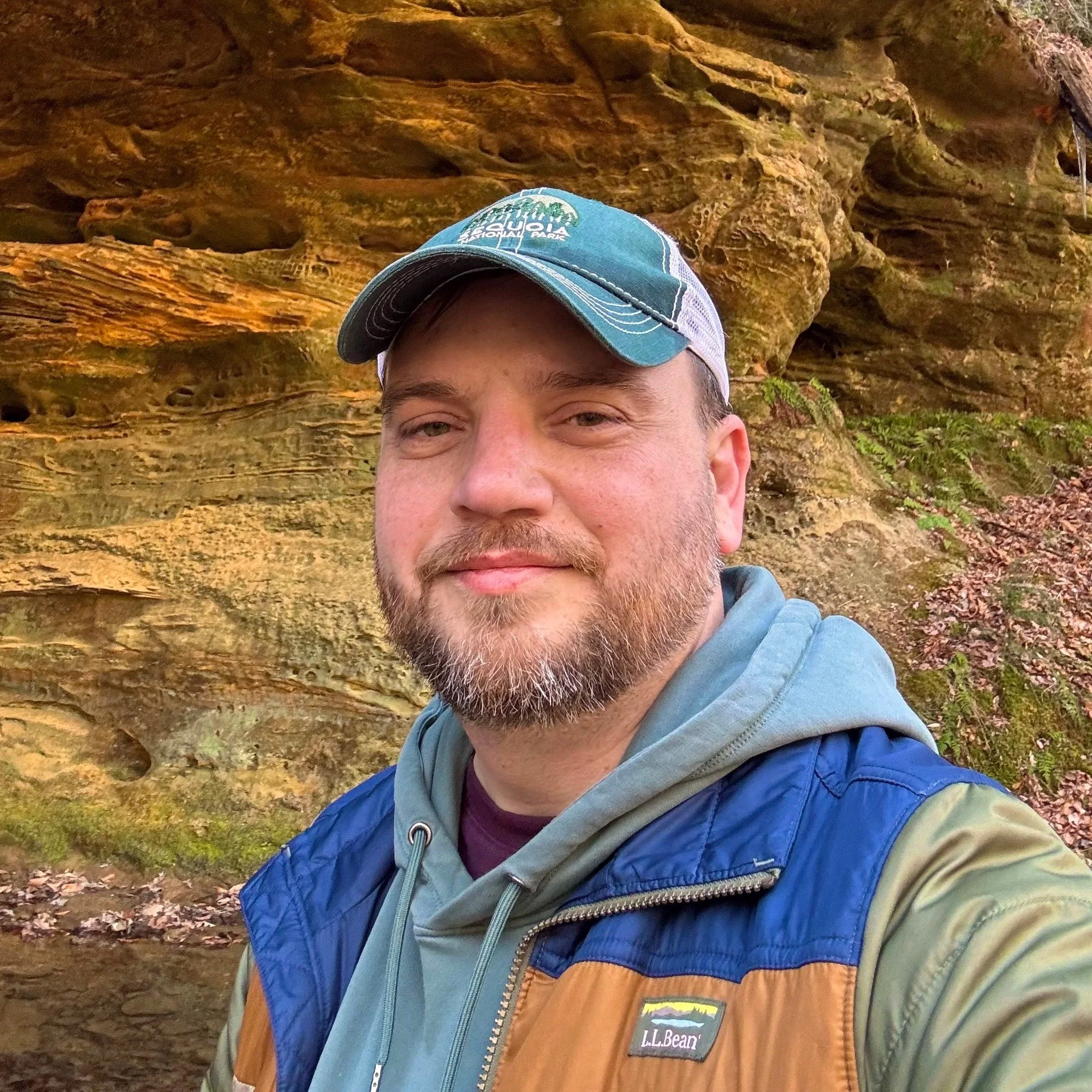 Partial view of a man's face with a beard and mustache, wearing a blue cap with a logo, standing in front of a rock formation with greenery.