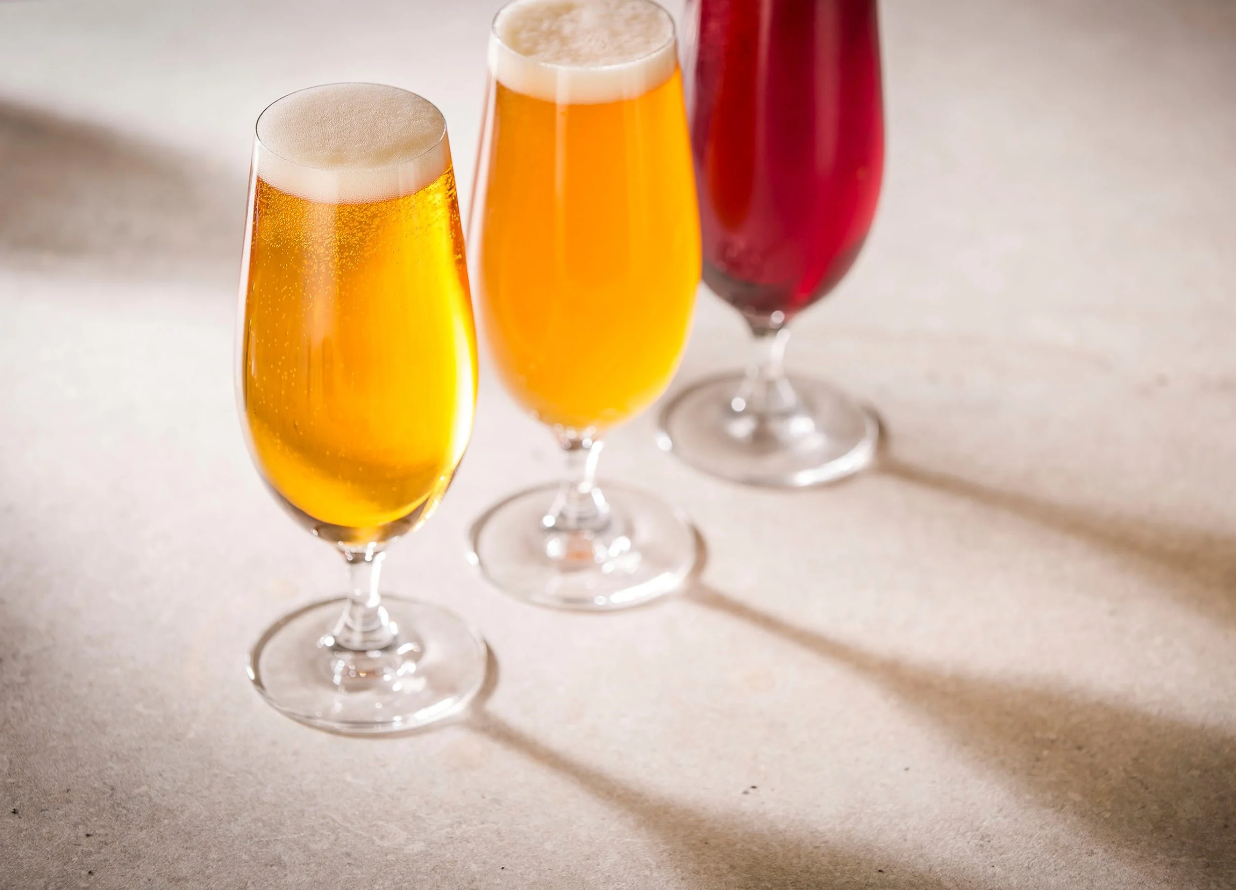 Three tulip glasses with foamy ciders in golden, orange, and red hues, casting long shadows on a light surface.