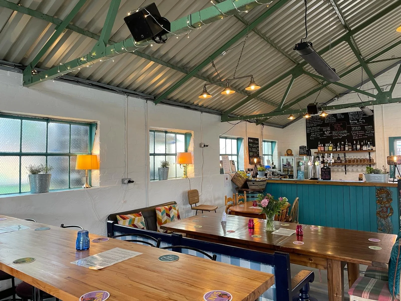 Napton Cidery's cozy, well-lit taproom with wooden tables, colorful cushions, and a teal bar counter; menu board in the background.
