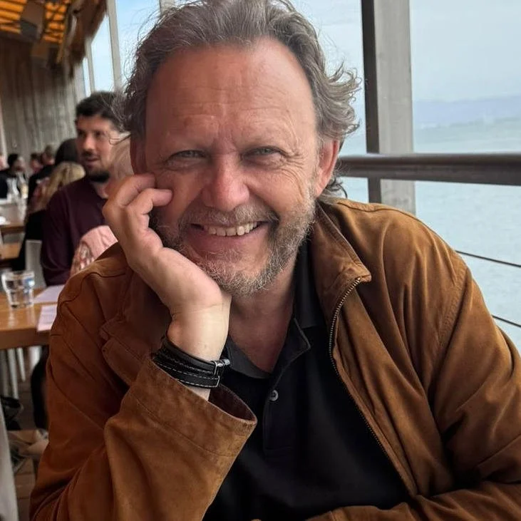 A smiling man with gray hair and a beard, wearing a brown jacket and black shirt, sitting at a restaurant with large windows and water view in the background.