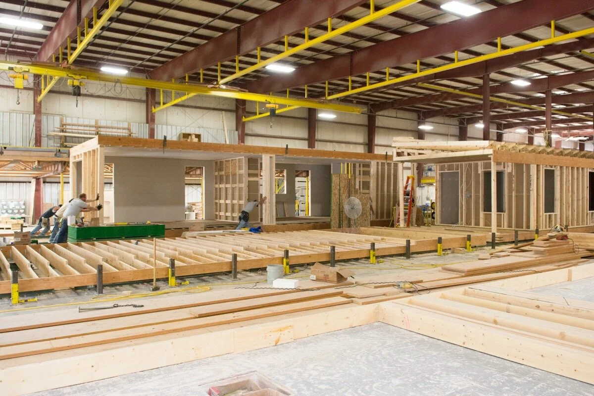 Construction workers building a wooden structure inside a large industrial warehouse, with several tools and materials around.
