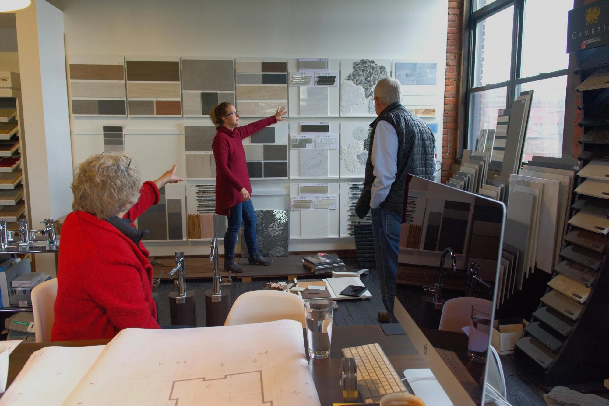 Interior of a tile and flooring store with three people discussing tile samples on display, a woman in a red sweater sitting at table points, a woman in a red jacket at the table, and a man in a black vest listening.