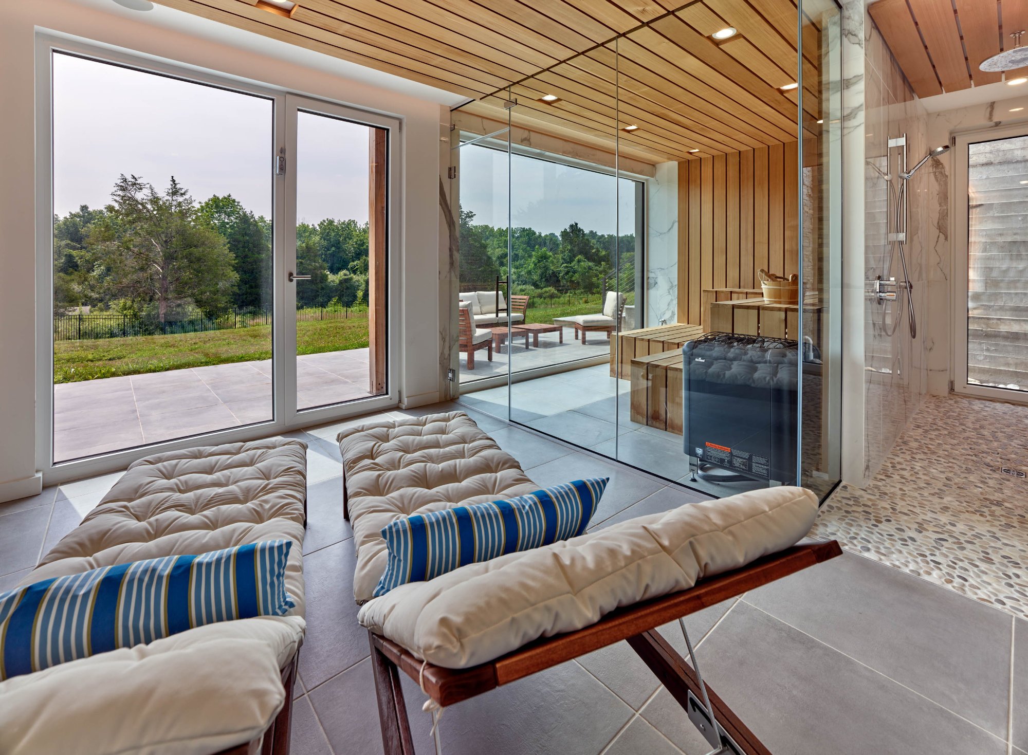 Contemporary indoor space with large glass doors and windows opening to an outdoor patio and green landscape, featuring seating areas and wooden accents.