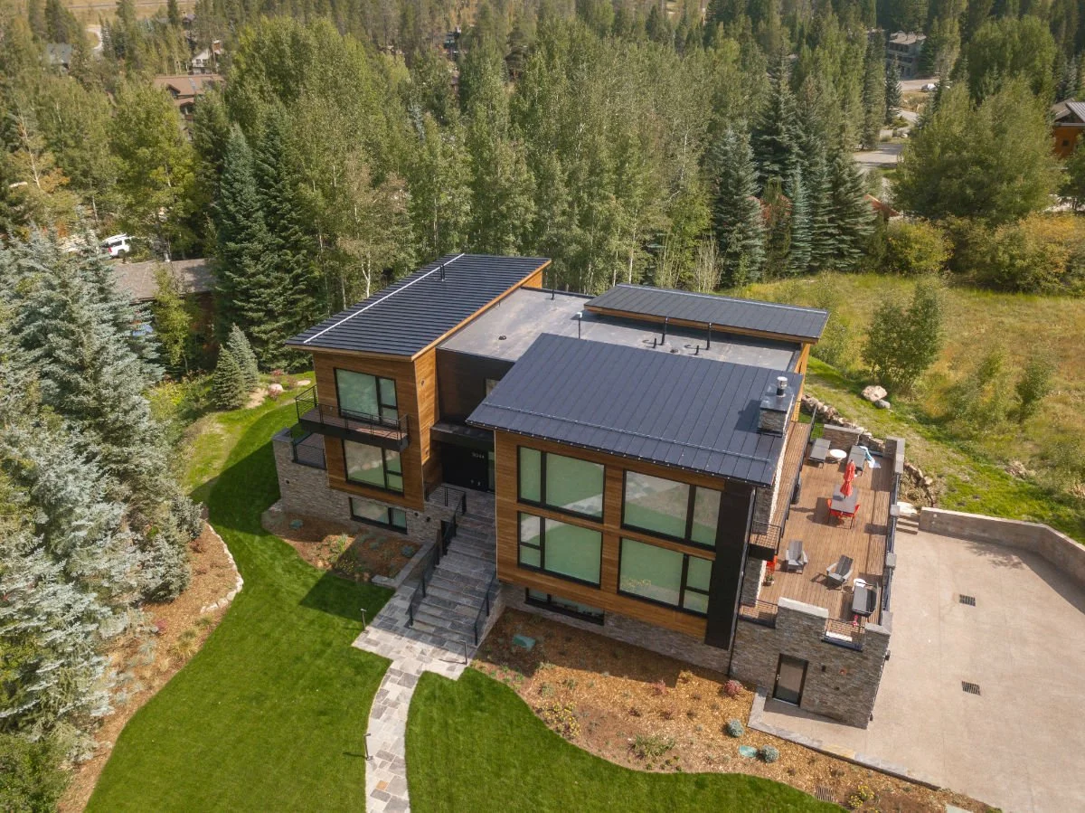 Aerial view of a modern house with flat and sloped roofs, large windows, a wooden deck with outdoor furniture and an umbrella, surrounded by trees and greenery.