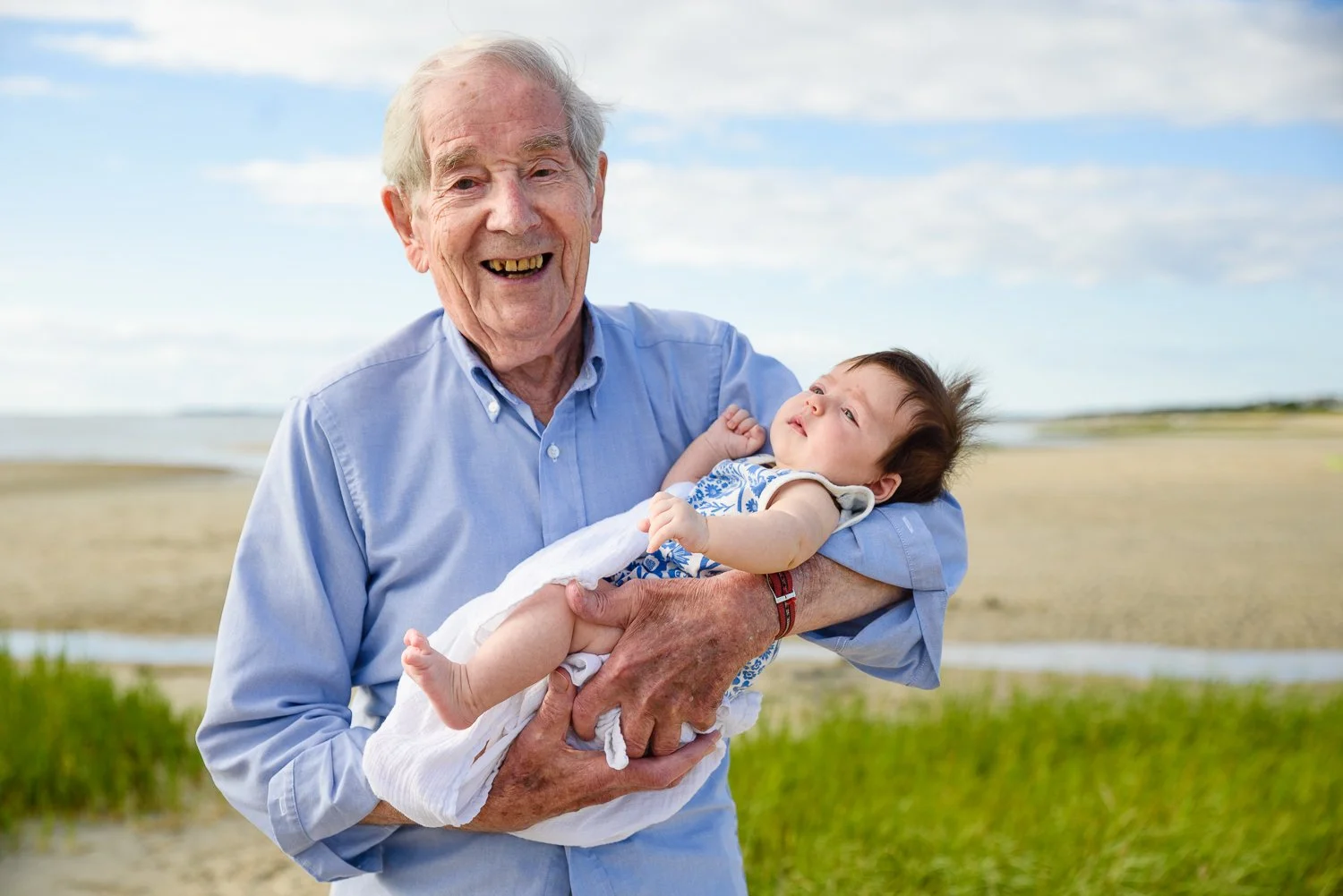 A man holds his infant great-grandaughter while standing on the beach. It's a sunny day, with blue sky & some clouds.