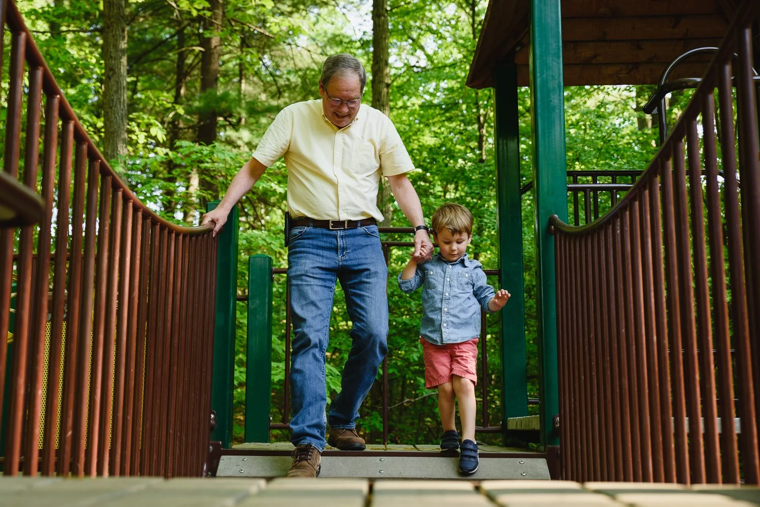 A grandfather and grandson cross a playground bridge in a park.