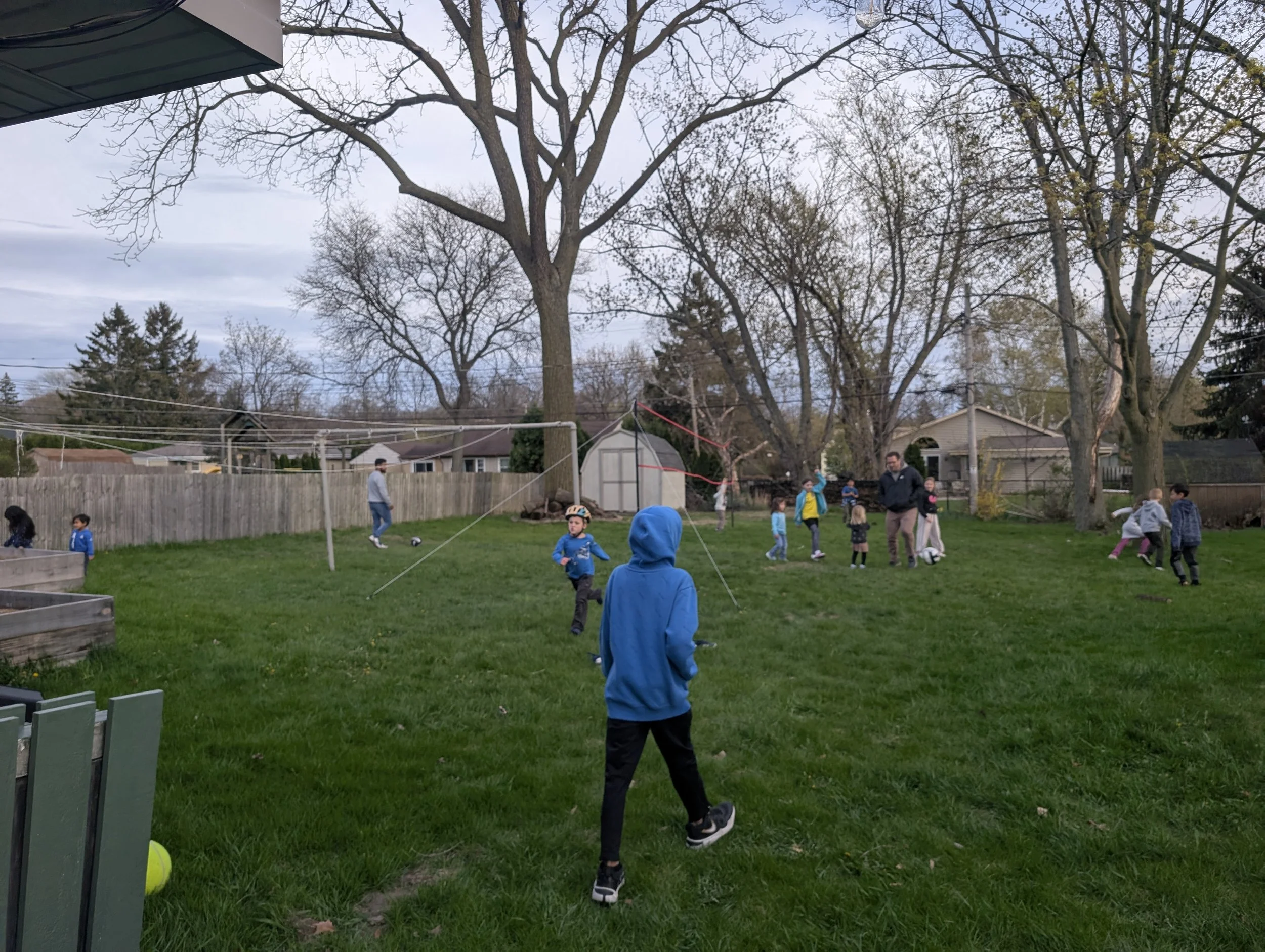 Kids playing volleyball in a backyard during overcast weather, with some children wearing helmets and jackets, and a large leafless tree, houses, and a shed in the background. They are participating in youth group at CrossWay Community Church