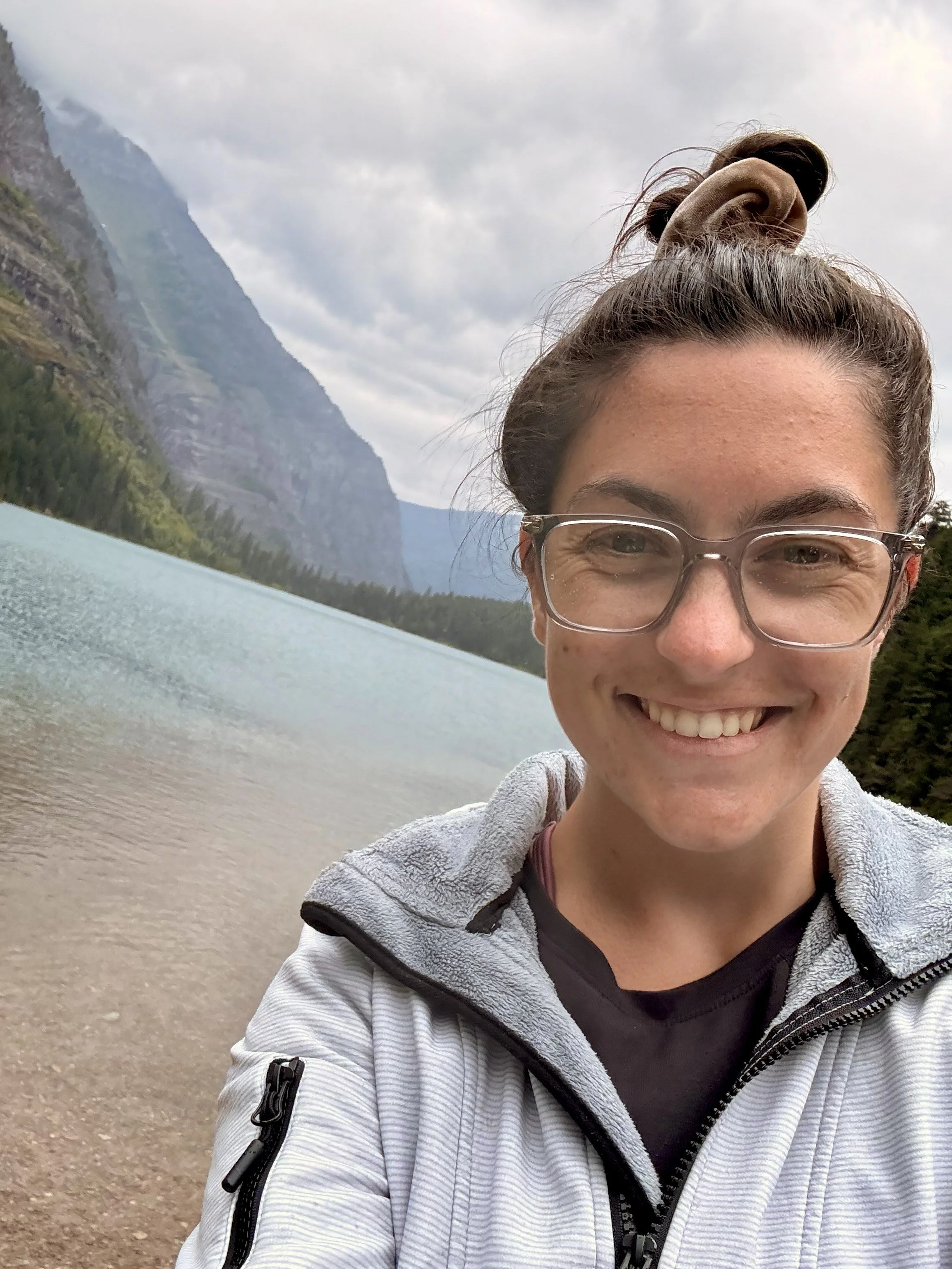 a woman stands with a scenic mountain background and is smiling at the camera. the woman has brown hair that is pulled into a bun, and is wearing glasses, a black shirt with a white zip-up jacket. the picture is cropped showing her face and the view