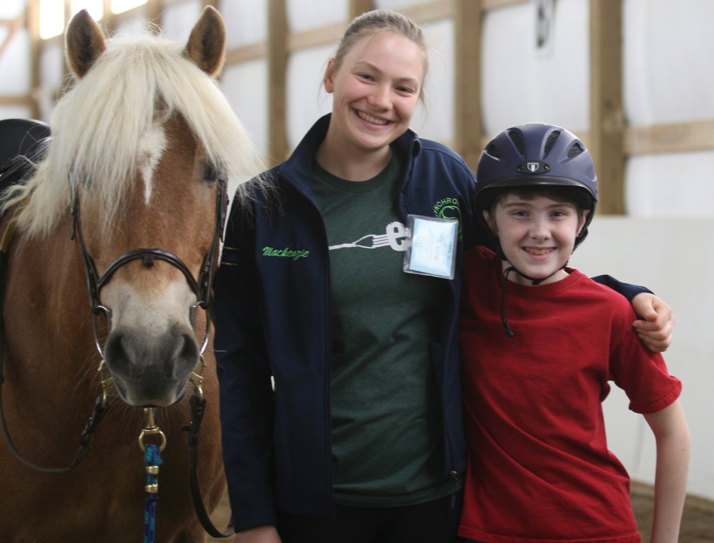   A white boy, wearing a helmet, stands next to a white woman who is standing next to a horse.  