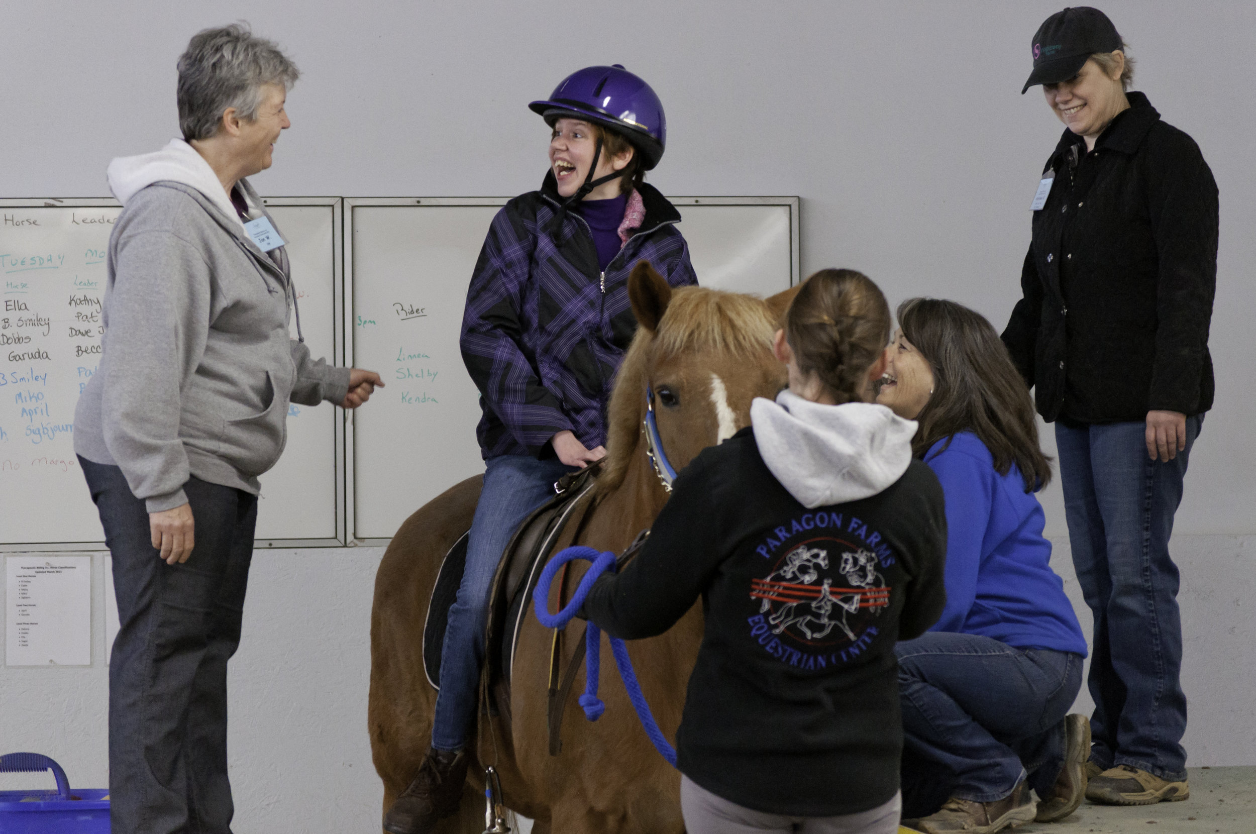   A white woman is on a horse. Three other white women, one on each side and one standing in front of the horse are looking at her. They are all smiling.  