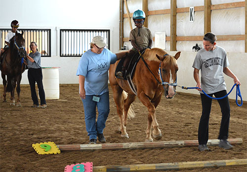   A black boy is riding a horse and is about to step over poles. A white woman walks next to him holding onto his leg. On the opposite side, a white woman leads the horse.  