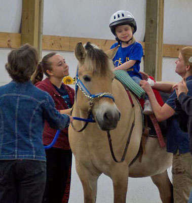   A young white girl is on a horse. People stand around her supporting her.  