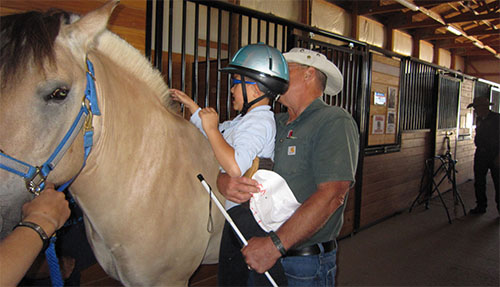   A white man holding a white cane holds a white boy up to pet a horse.  