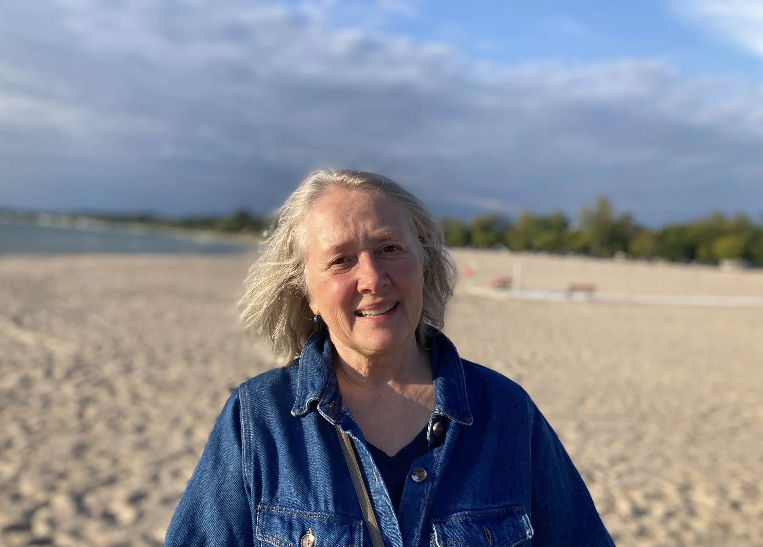 an older woman with light grey hair stands on the beach smiling at the camera. the woman is wearing a blue jean jacket.