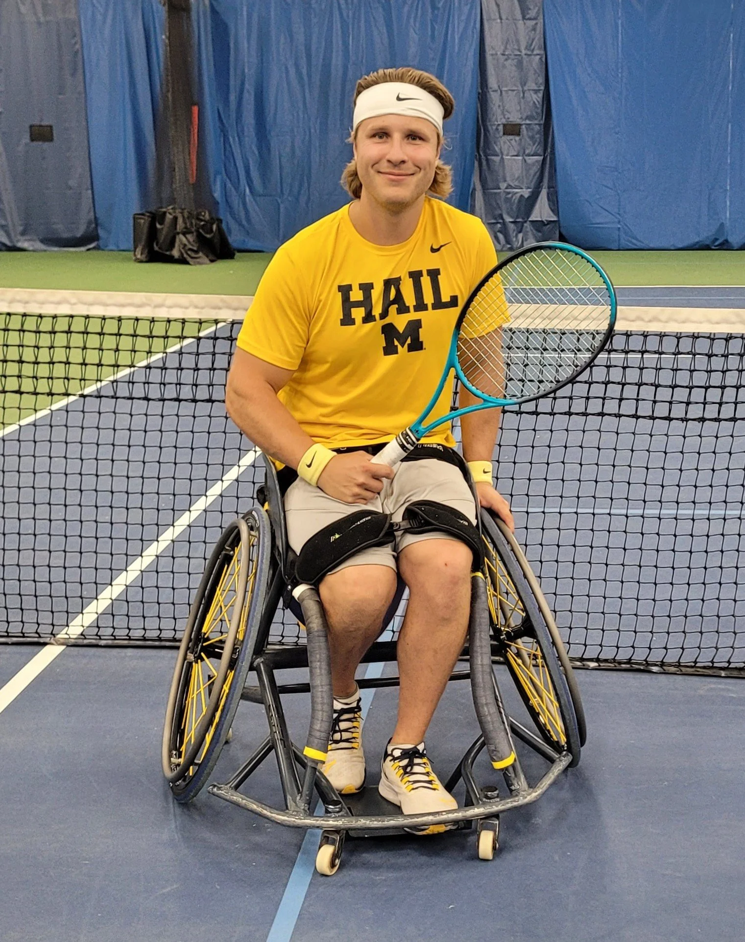 the man pictured has brown hair, is wearing a white Nike sweatband, a yellow UofM shirt, and black shorts. the man is in a wheelchair and is smiling.