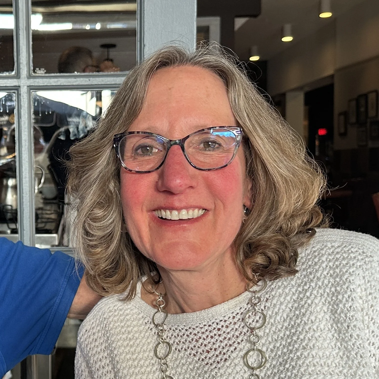 a woman with darker blonde hair is smiling at the camera. the woman is waring darker colored glasses, a white sweater, and a necklace.