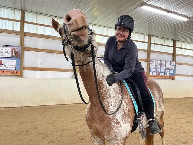 a woman sitting on a tan horse smiling. the woman is wearing a black helmet, glasses, black long sleeved shirt, black pants, and boots.