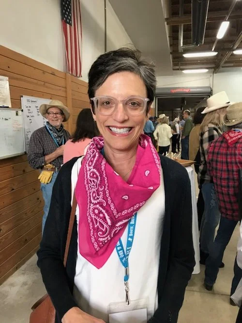 a woman with darker hair is smiling and looking at the camera. the woman is wearing tan glasses, a black shirt with a white vest, and a colorful red western scarf.