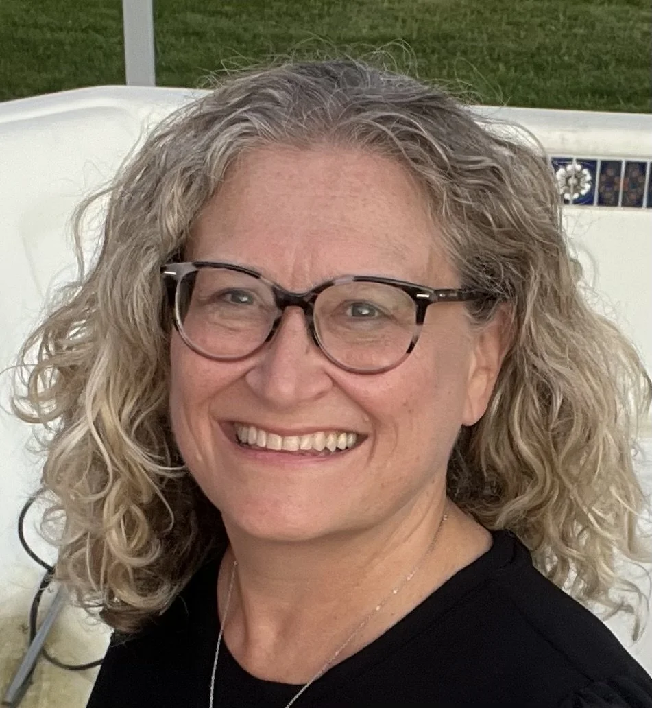 a woman with light brown, curly hair smiles at the camera. she is wearing black glasses, a black shirt, and silver jewelry.