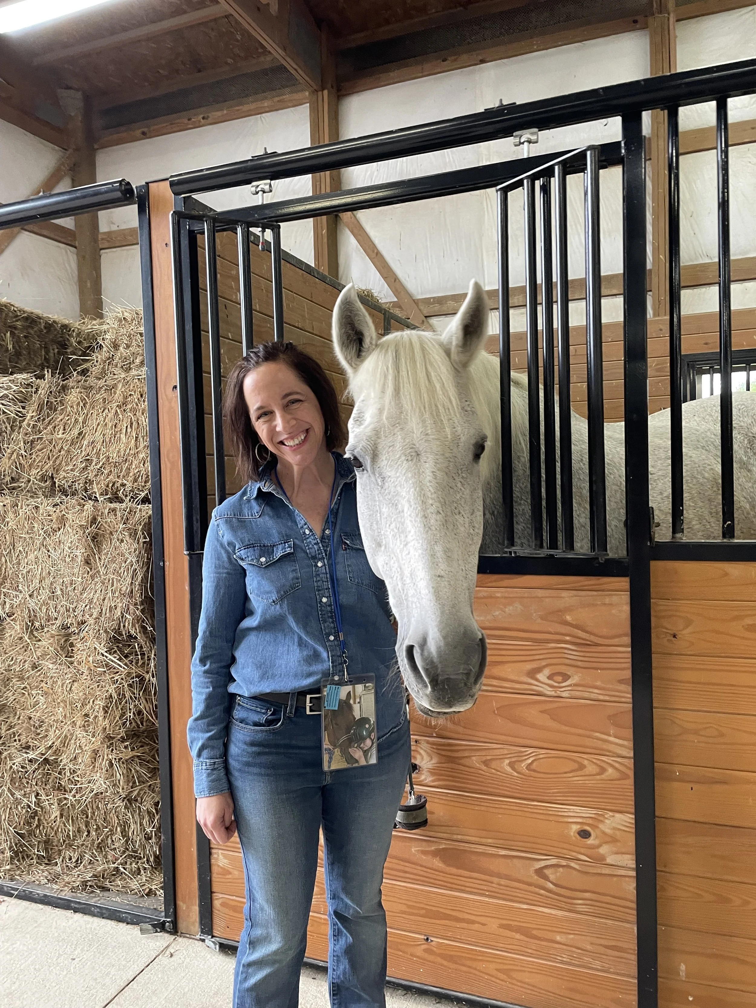 a woman stands next to a white horse in a stall. the woman wears a blue jean shirt and blue jeans. she has shoulder length brown hair and is smiling.